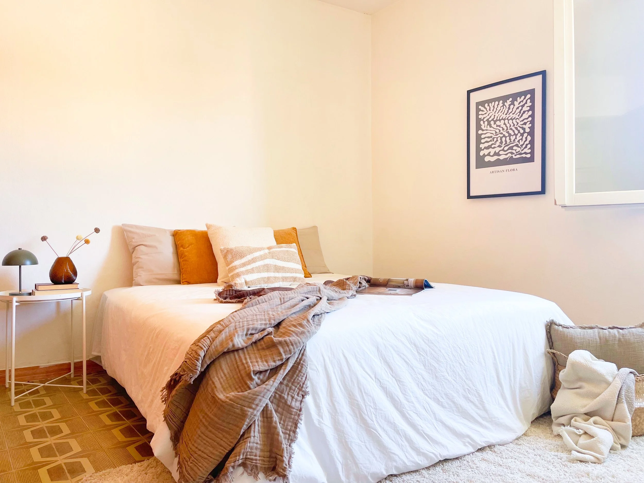 Camera da letto luminosa con letto matrimoniale, copriletto bianco, cuscini colorati, lampada e libri su un tavolino, poster sul muro.