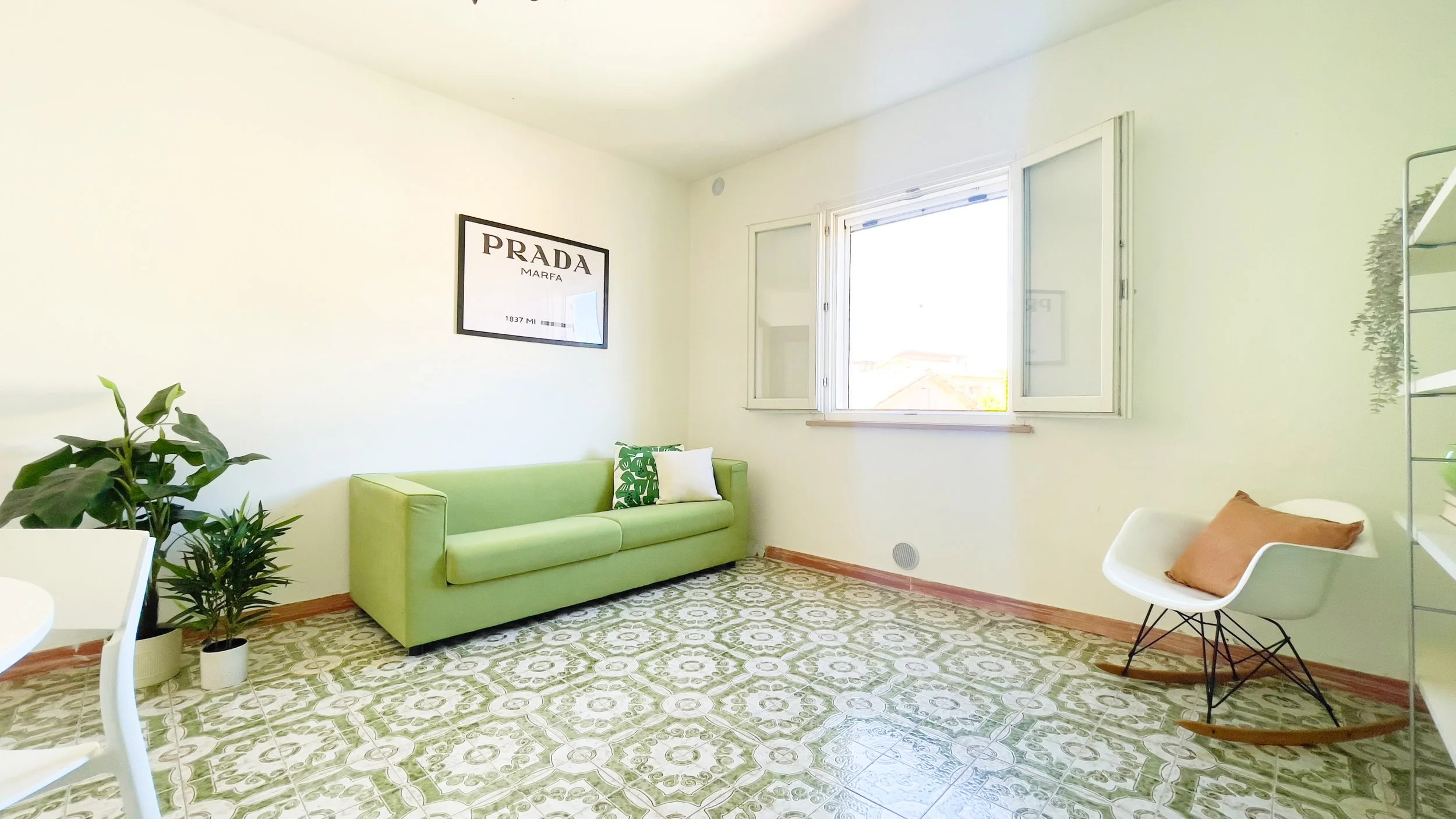 Soggiorno luminoso con divano verde, sedia a dondolo bianca, pianta in vaso, e poster sulla parete. Pavimento con piastrelle motivo geometrico.