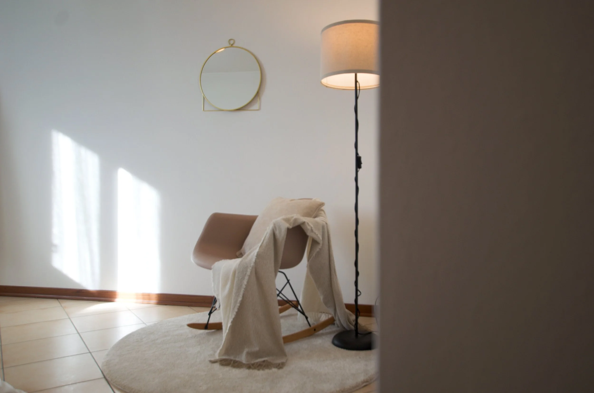 Interno di una stanza con una sedia a dondolo moderna coperta da una coperta beige, una lampada da terra e uno specchio rotondo appeso al muro bianco.