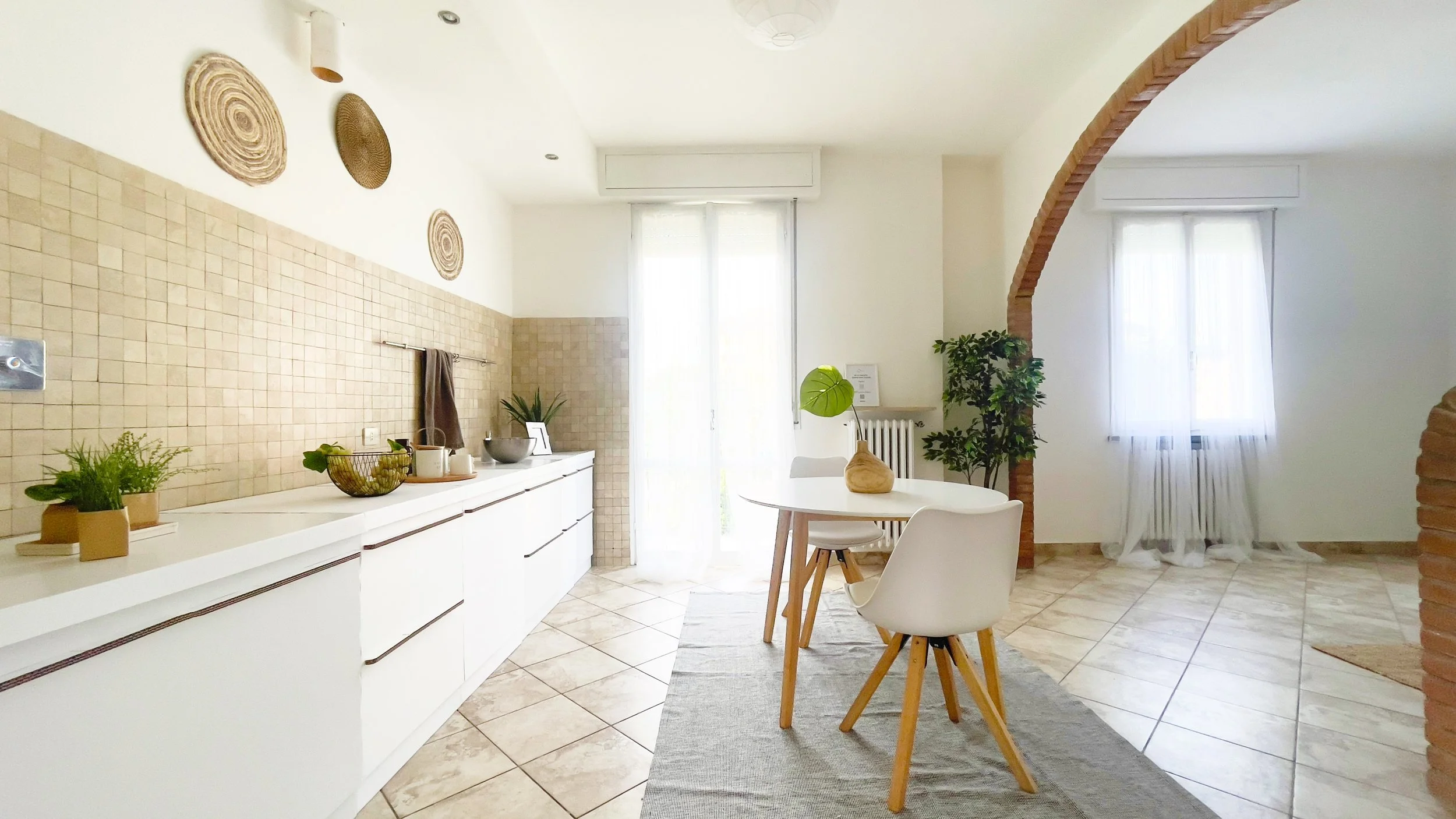 Cucina moderna con mobili bianchi, tavolo rotondo, sedie bianche, pianta ornamentale, decorazioni murali in vimini, arco in mattoni e finestre con tende bianche.