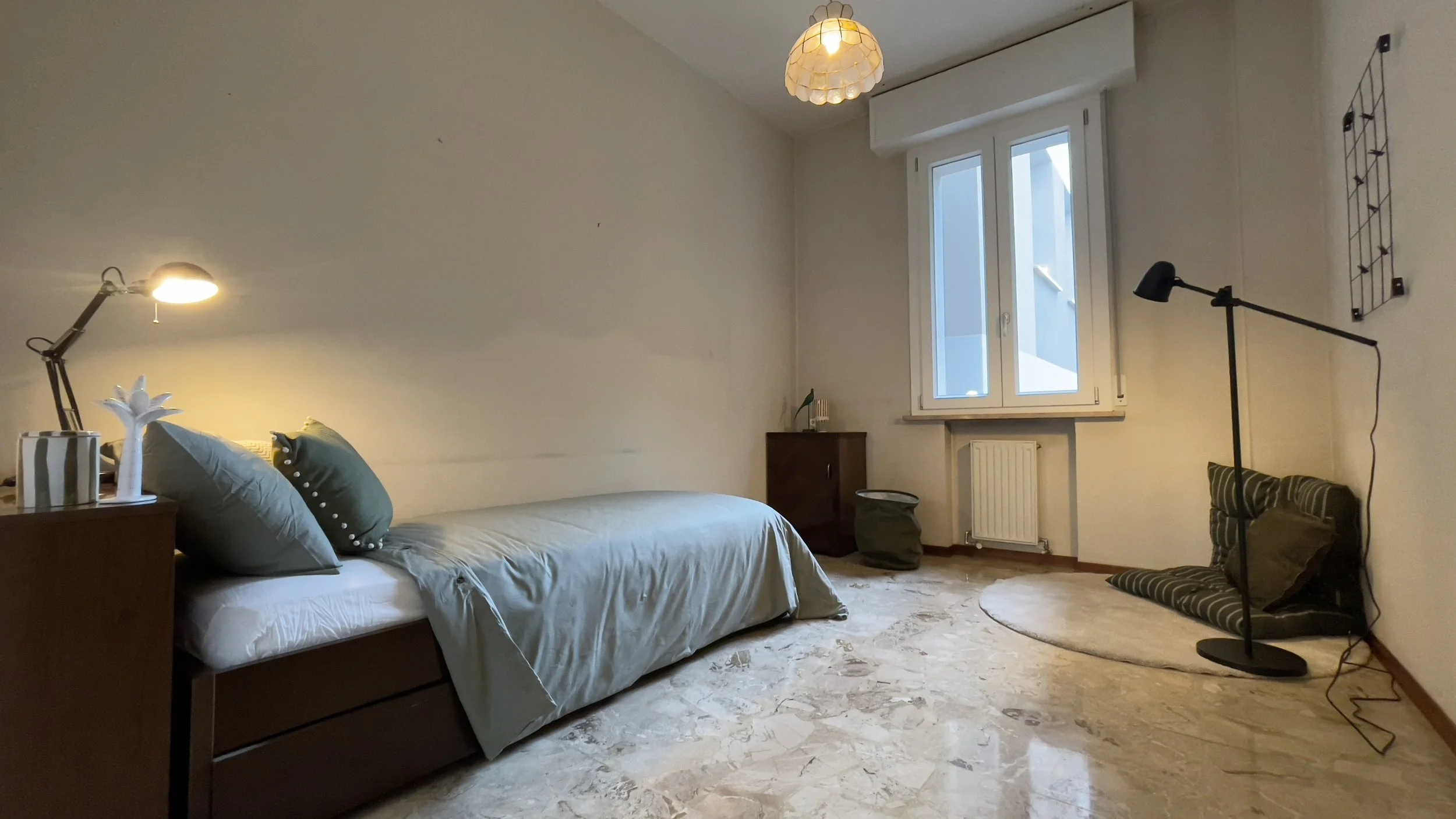 Camera da letto semplice con letto singolo, lampada da lettura, finestra, lampada a piantana e cuscini sul pavimento.