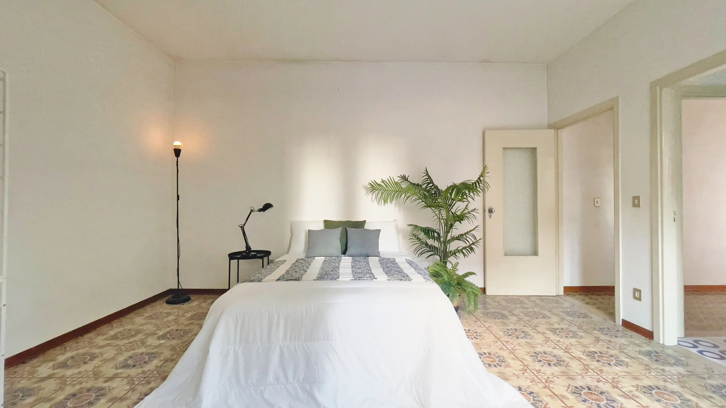 Camera da letto minimalista con letto matrimoniale, lampada da terra, lampada da tavolo nera su comodino, pianta verde accanto al letto, porta bianca e pavimento piastrellato.