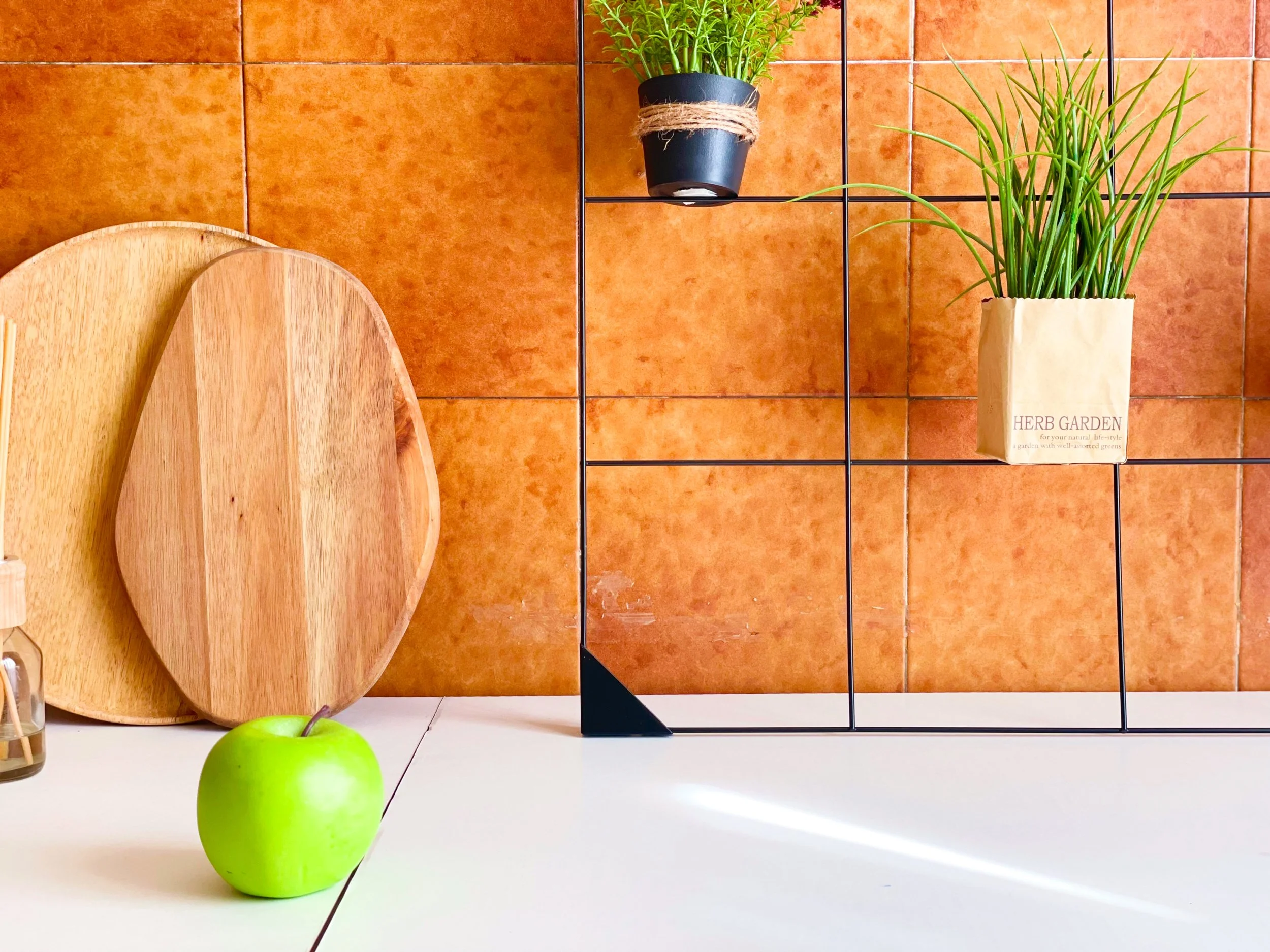 Cucina con sfondo di piastrelle arancioni, tavolino bianco, due taglieri di legno sovrapposti, una mela verde, una griglia metallica con piante in vaso.