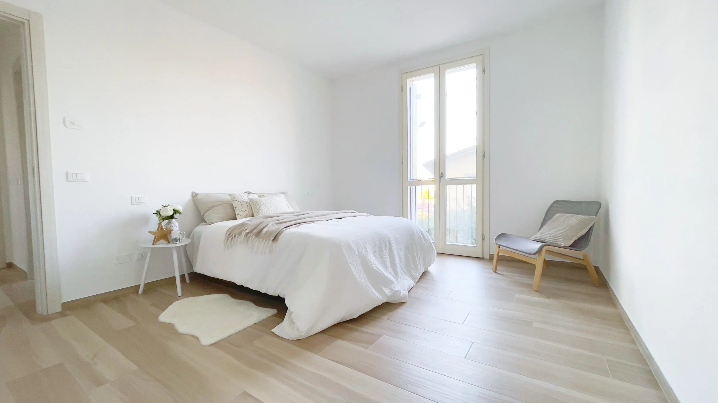 Camera da letto minimalista con letto matrimoniale, biancheria bianca, comodino con decorazioni, sedia con cuscino, pavimento in legno chiaro e finestra luminosa.