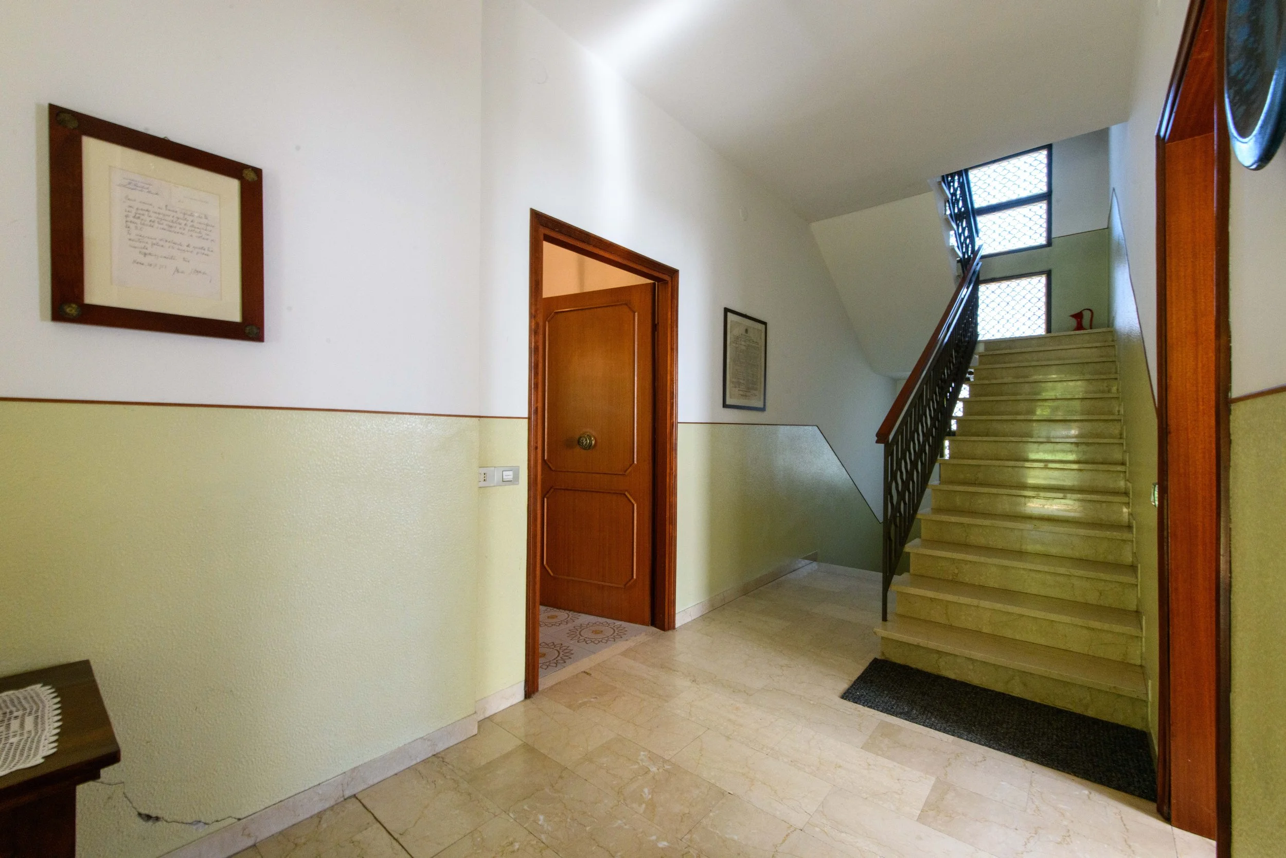 Ingresso di un edificio con scale in marmo, porta di legno e quadri appesi alle pareti.