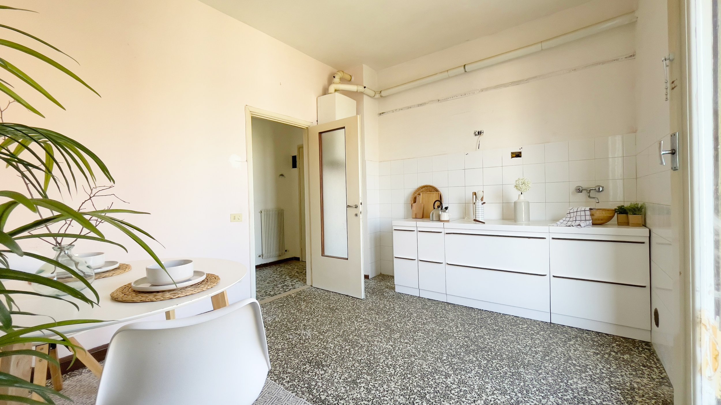 Cucina moderna luminosa con mobili bianchi, tavolo rotondo con piatti, piante verdi, pavimento in terrazzo e pareti beige chiaro.