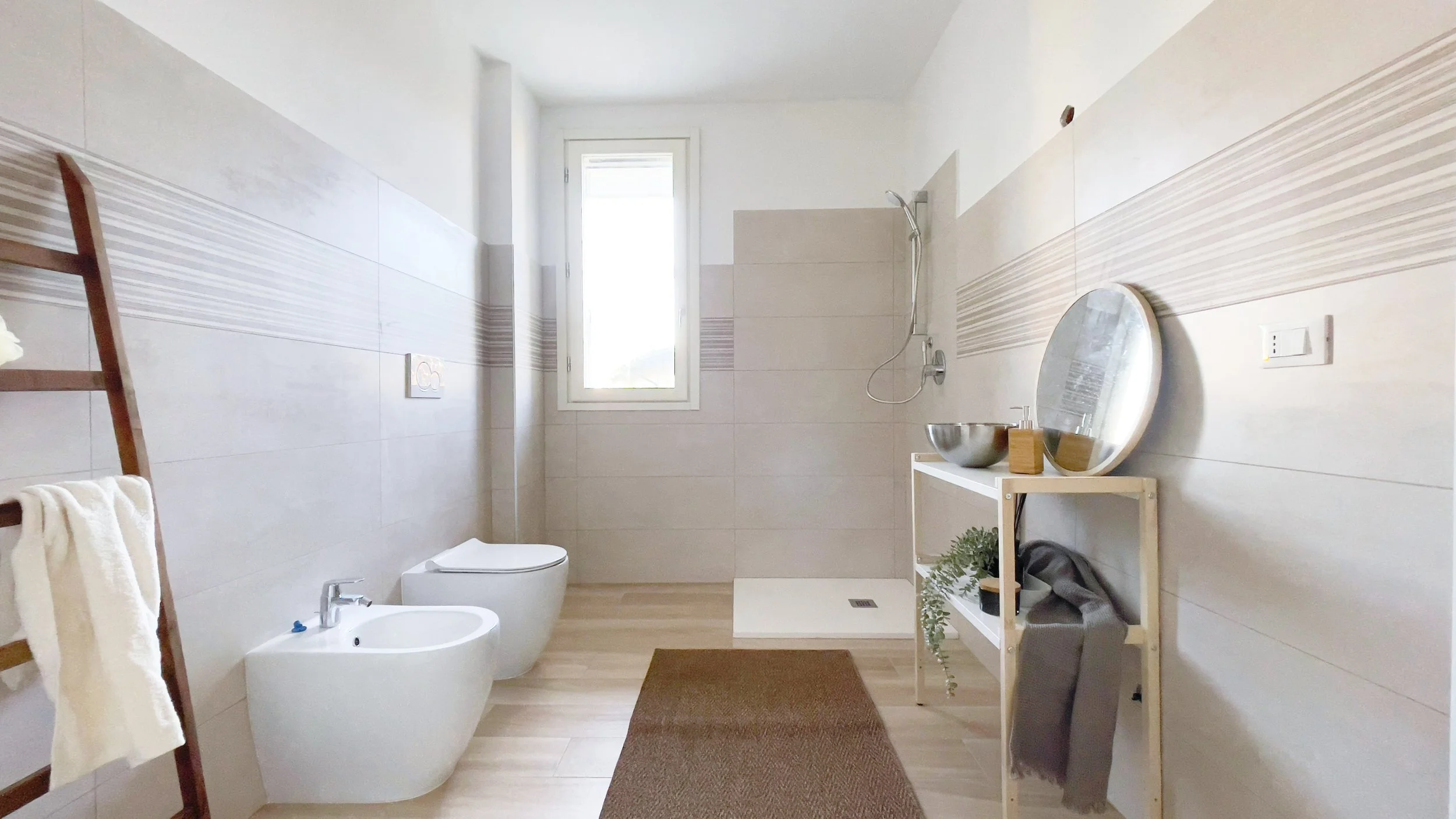 Bagno moderno con sanitari bianchi, una doccia, una finestra, una scala porta asciugamani, e un mobile con specchio e decorazioni.