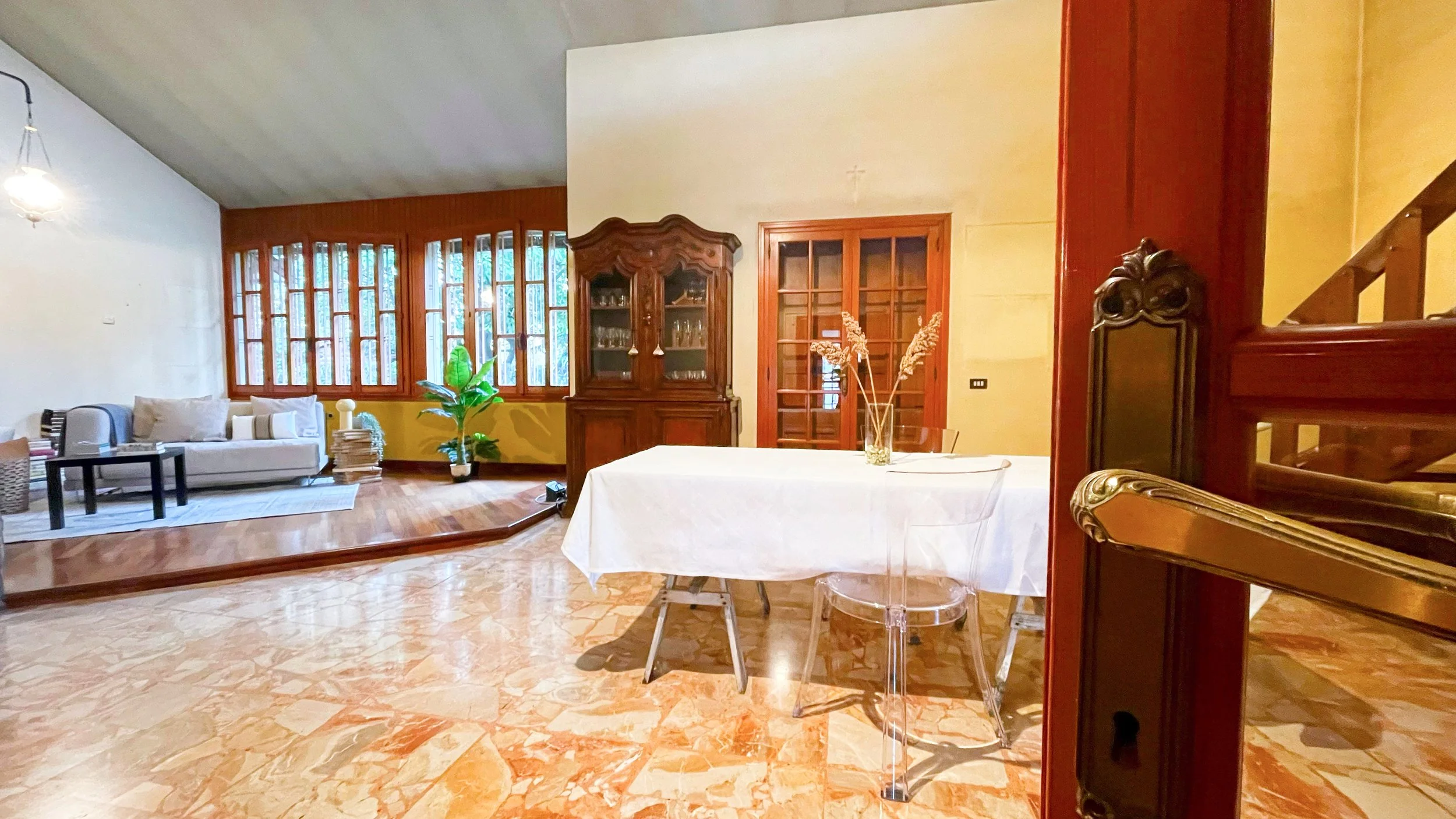 Soggiorno con pavimento in marmo, tavolo con tovaglia bianca, sedie trasparenti, mobile in legno e divano con cuscini, pianta decorativa e lampadario.