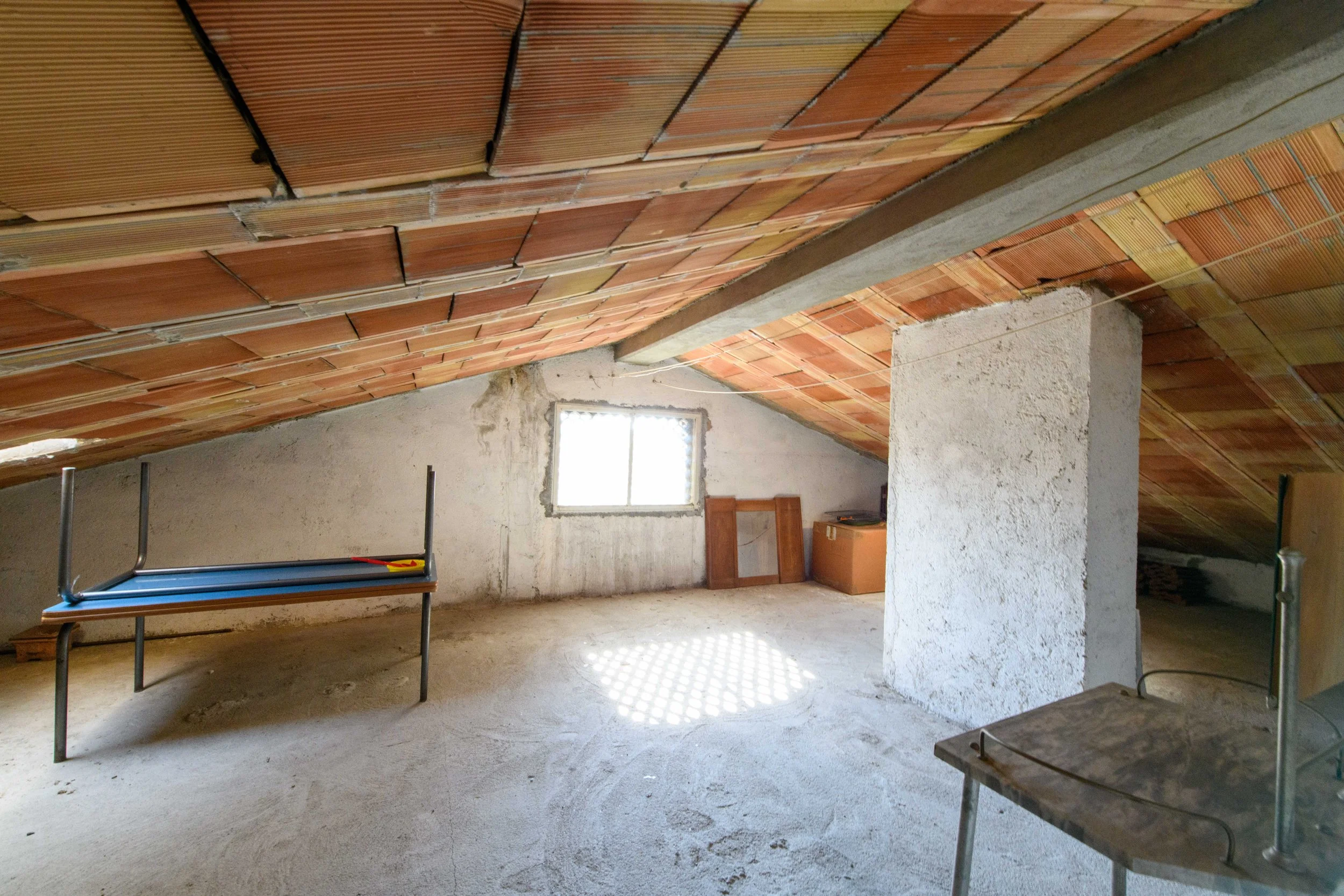 Soffitta spaziosa con tetto in tegole, finestra piccola, tavolo capovolto, pareti bianche e scatole di cartone.