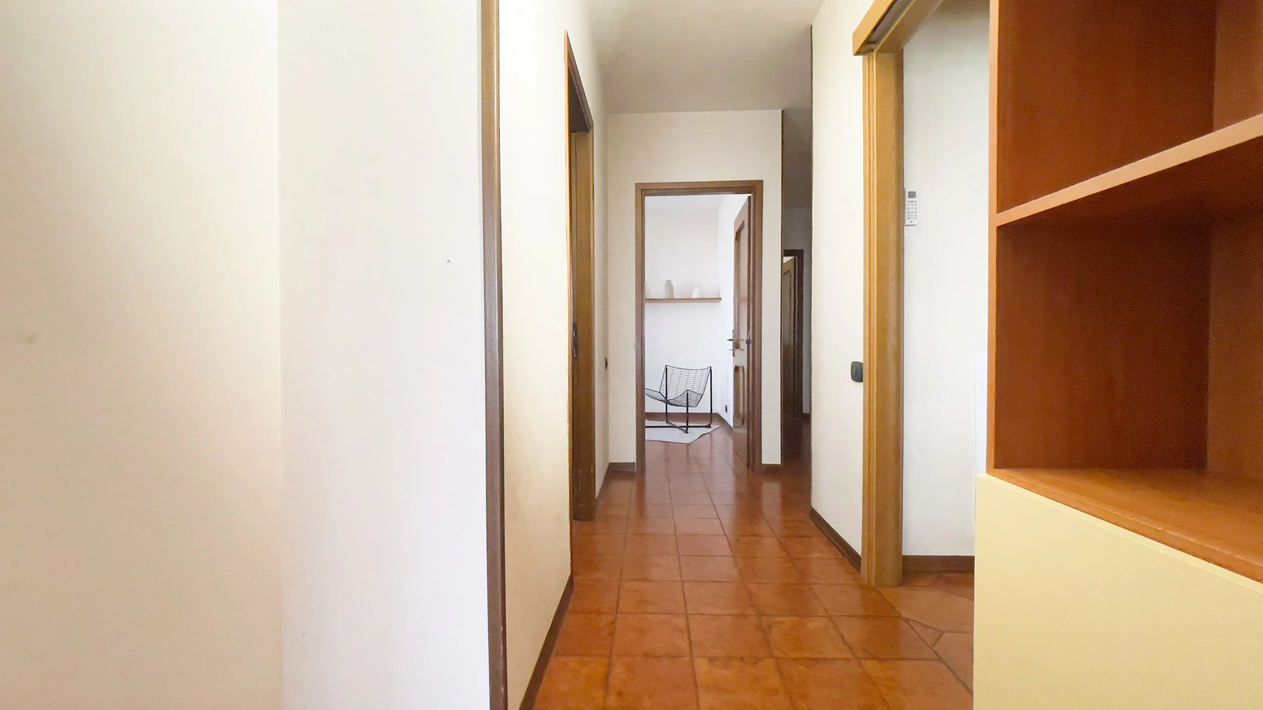 Corridoio di una casa con pavimento in piastrelle arancioni, pareti bianche e una libreria in legno sulla destra. Due porte aperte sul lato destro conducono a stanze diverse. Alla fine del corridoio c'è una sedia moderna di design e un piccolo tappet