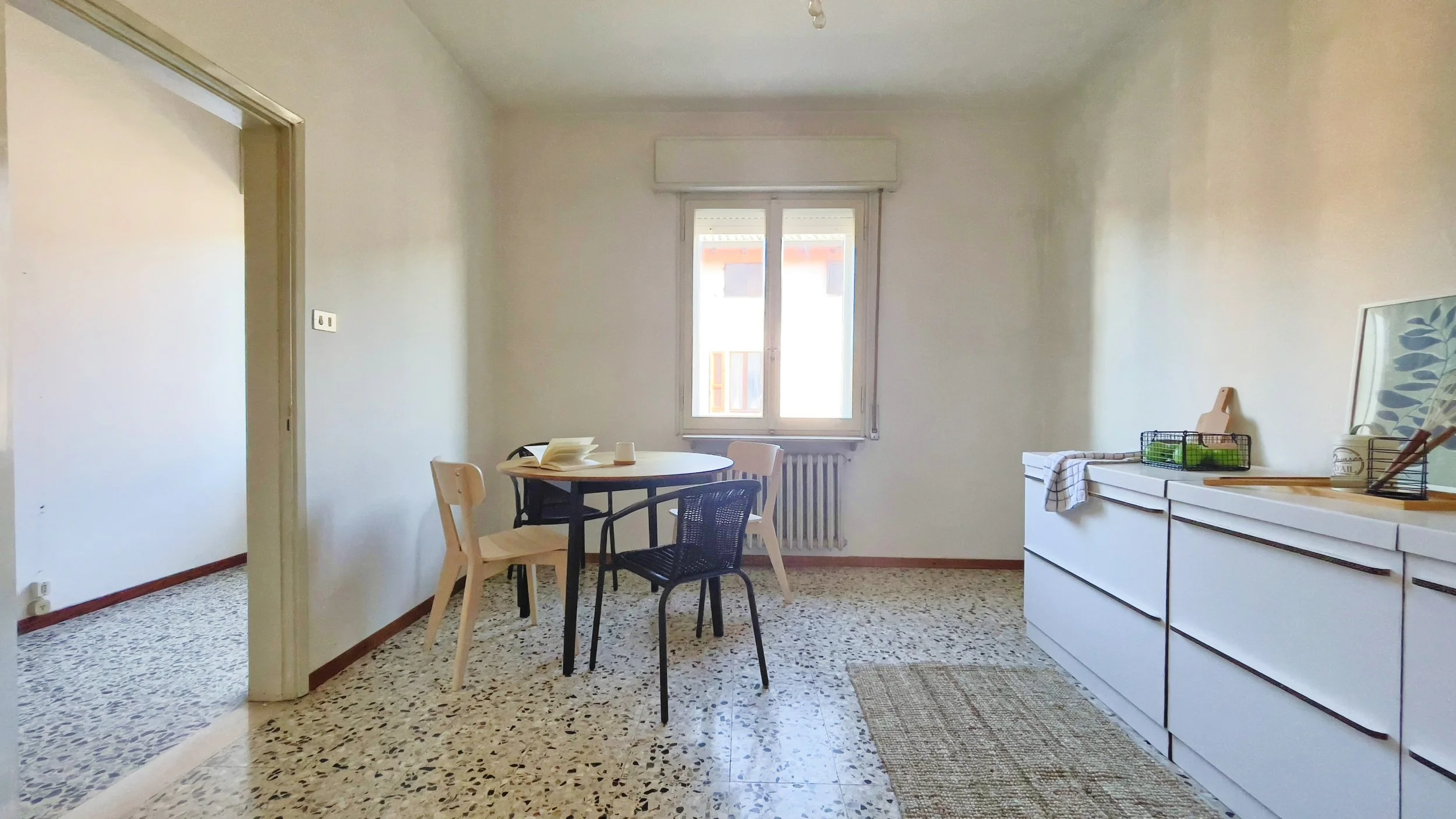 Interno di una stanza luminosa con un tavolo rotondo e sedie, pavimento in graniglia, una finestra grande e una credenza con oggetti decorativi.