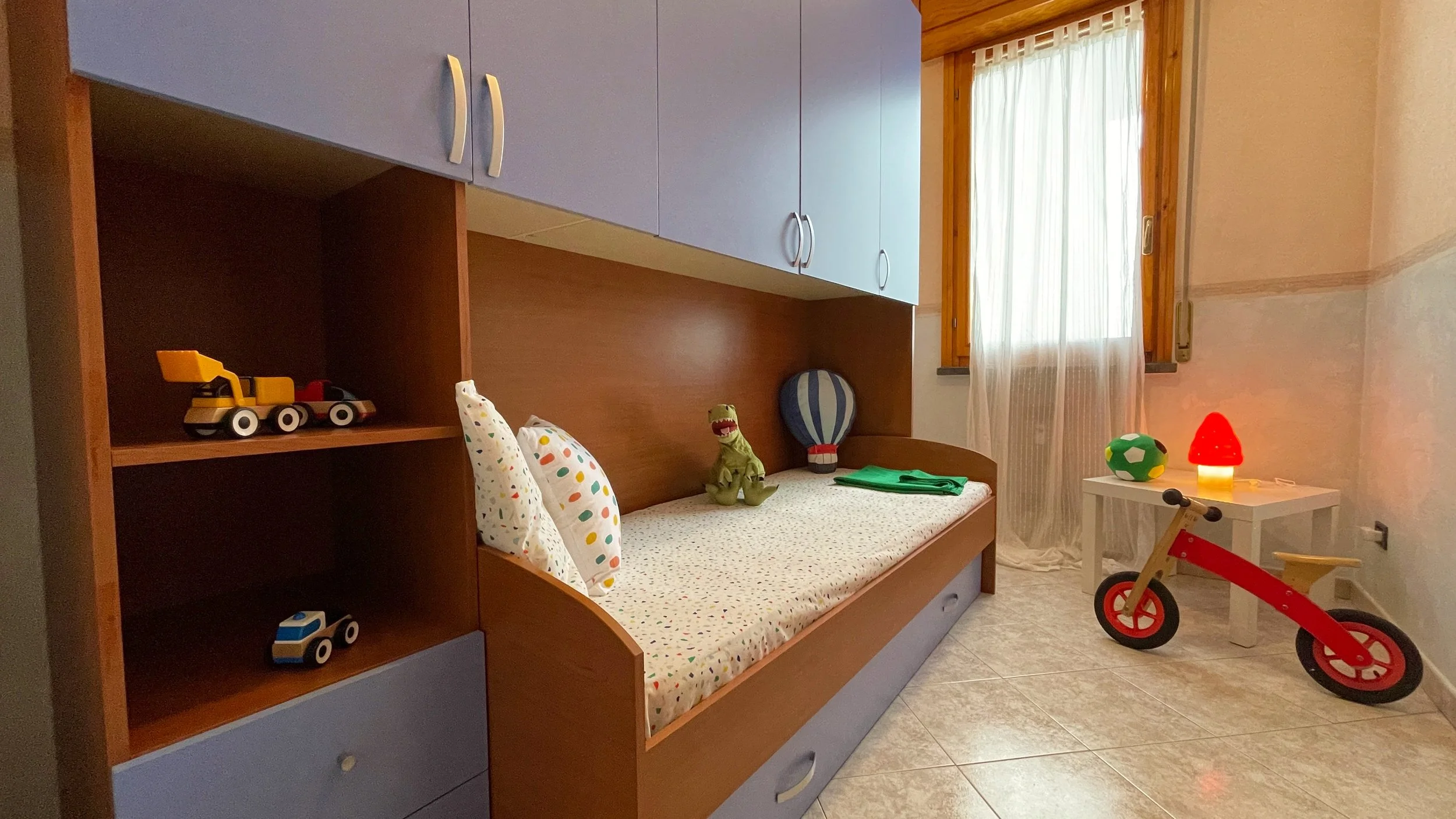 Camera da letto per bambini con letto singolo, scaffali con giocattoli, finestra con tenda bianca, lampada a forma di fungo rosso, e bicicletta senza pedali rossa.