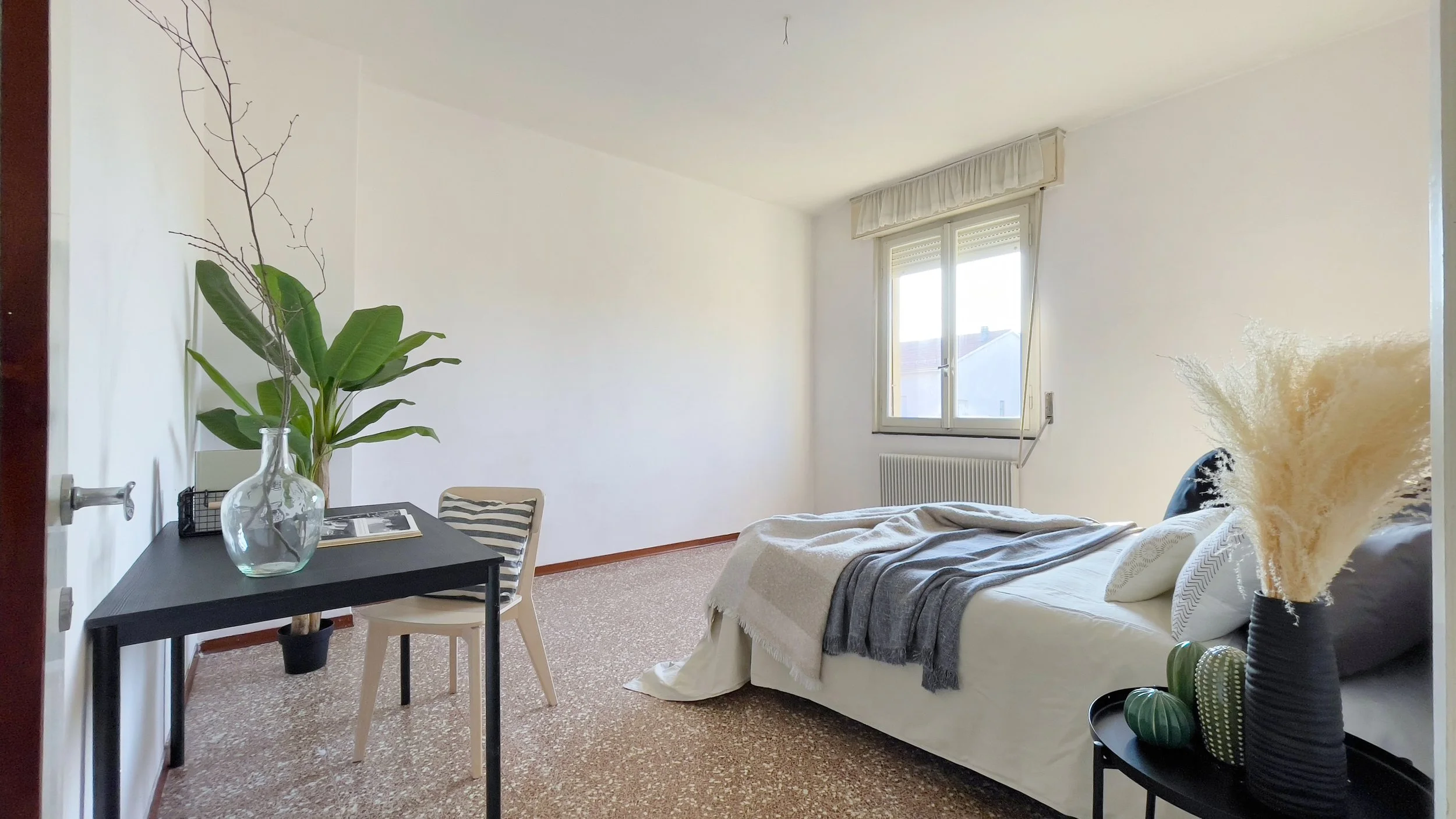 Camera da letto minimalista con letto, scrivania nera, sedia, piante decorative e una finestra.