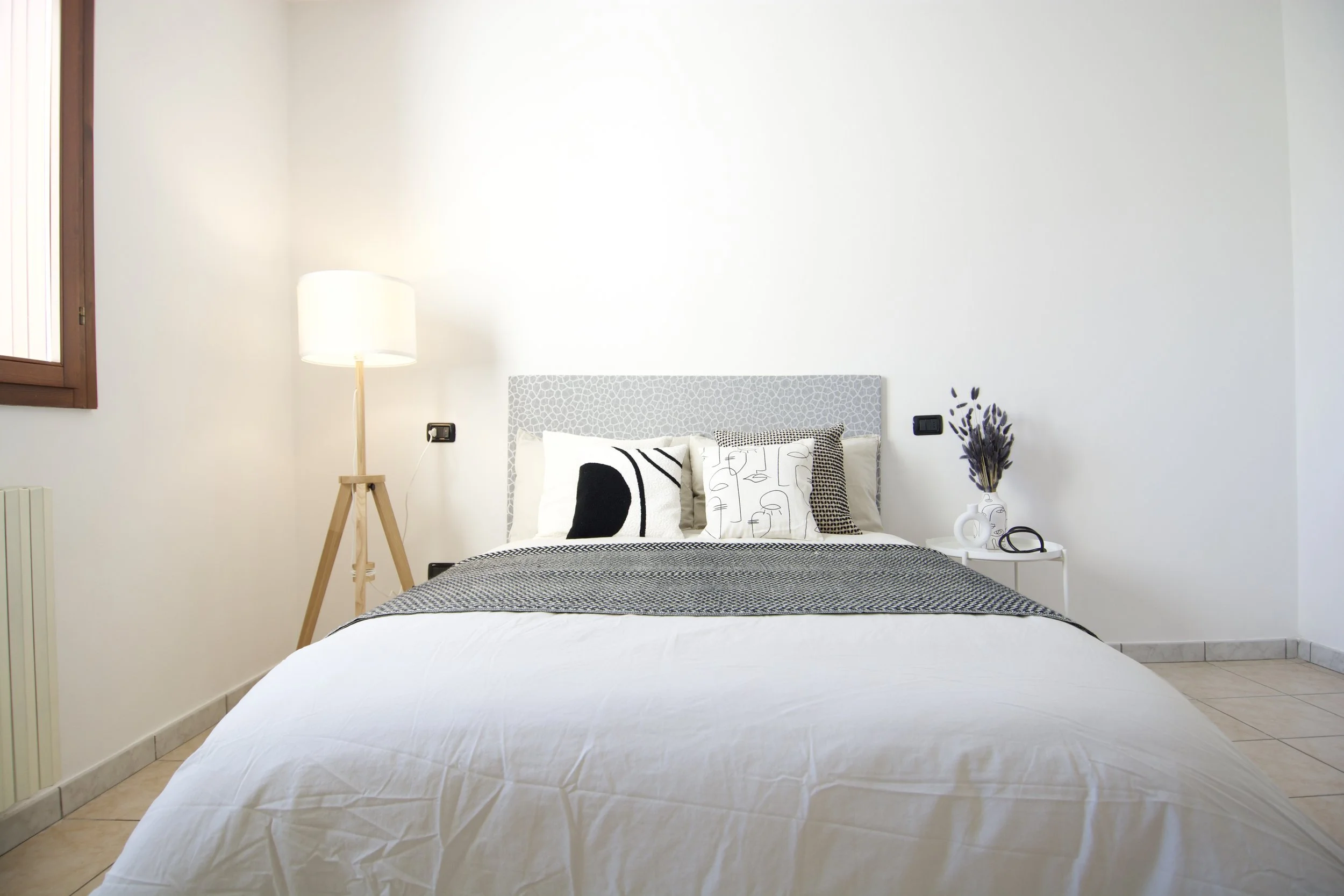 Camera da letto moderna con letto matrimoniale, cuscini decorativi, lampada da terra, finestra, e comodino con vaso e occhiali.