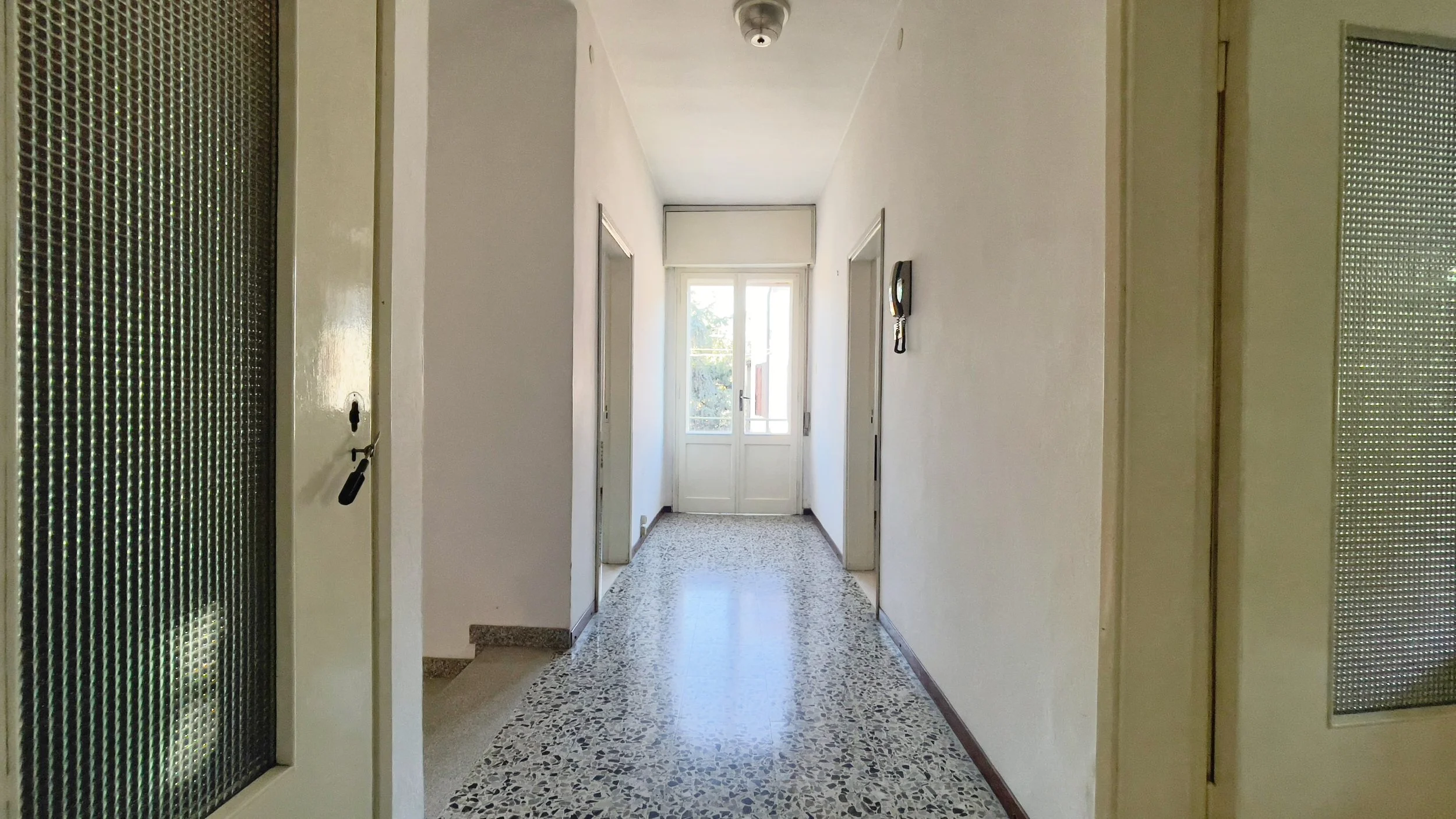 Corridoio di una casa con porte bianche e pavimento in graniglia, al fondo una porta finestra che lascia entrare la luce.