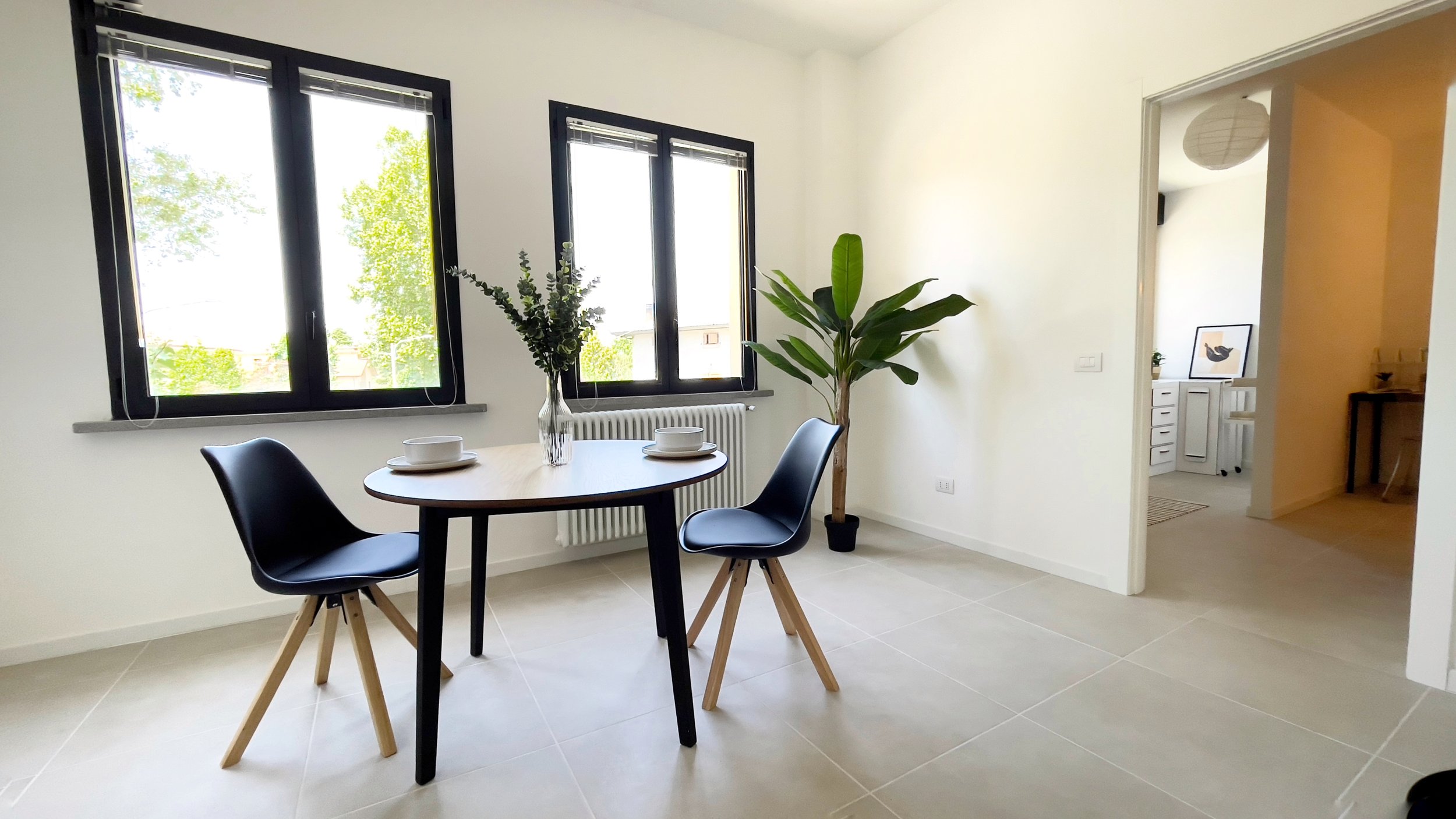 Sala da pranzo moderna con tavolo rotondo, due sedie nere, pianta in vaso, grandi finestre e vista su stanza adiacente. Pareti bianche e pavimento piastrellato.