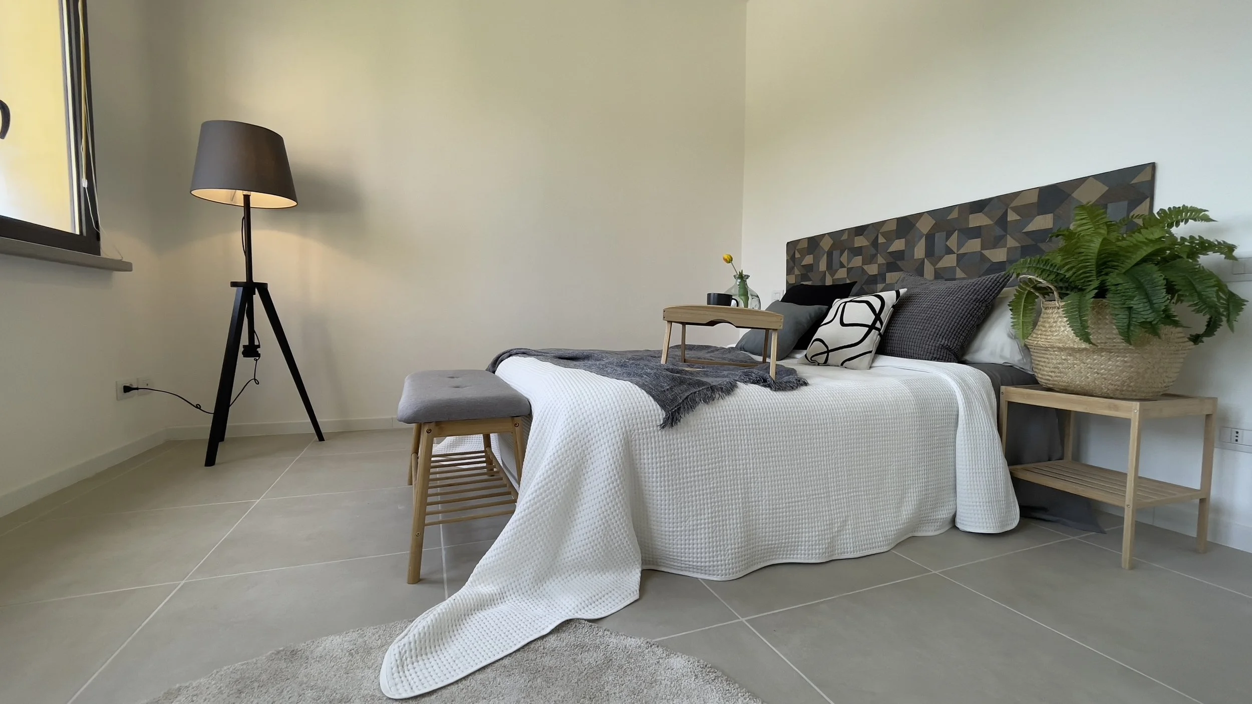 Camera da letto moderna con letto matrimoniale, coperta bianca, cuscini decorativi, abat-jour a treppiede nero, panca di legno, vaso con fiore giallo e pianta in vaso di vimini su un comodino.