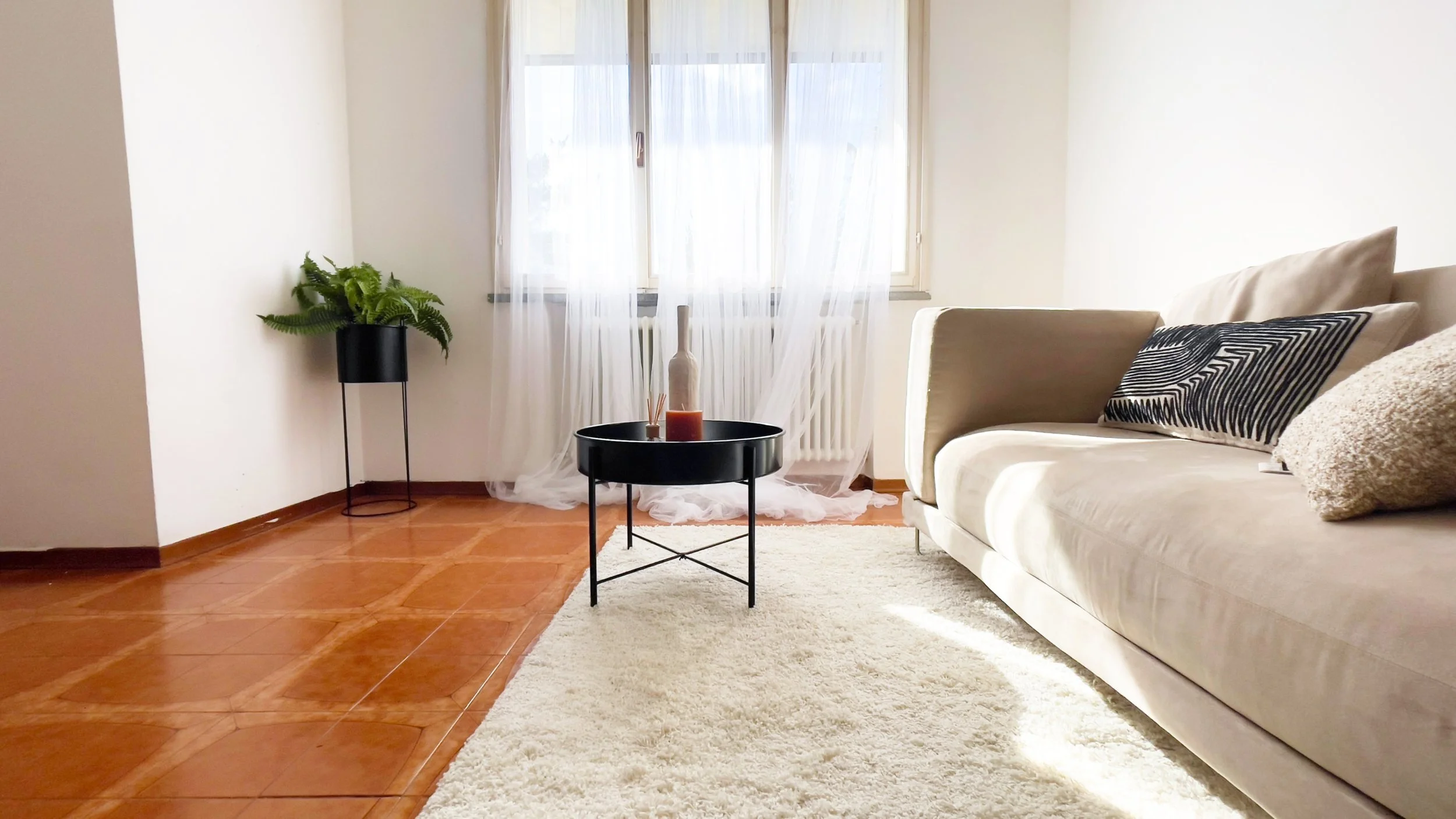 Soggiorno moderno con divano beige, cuscini decorativi, tavolino nero con candele, tappeto bianco e pianta sul lato sinistro. La finestra con tende trasparenti lascia entrare luce naturale.