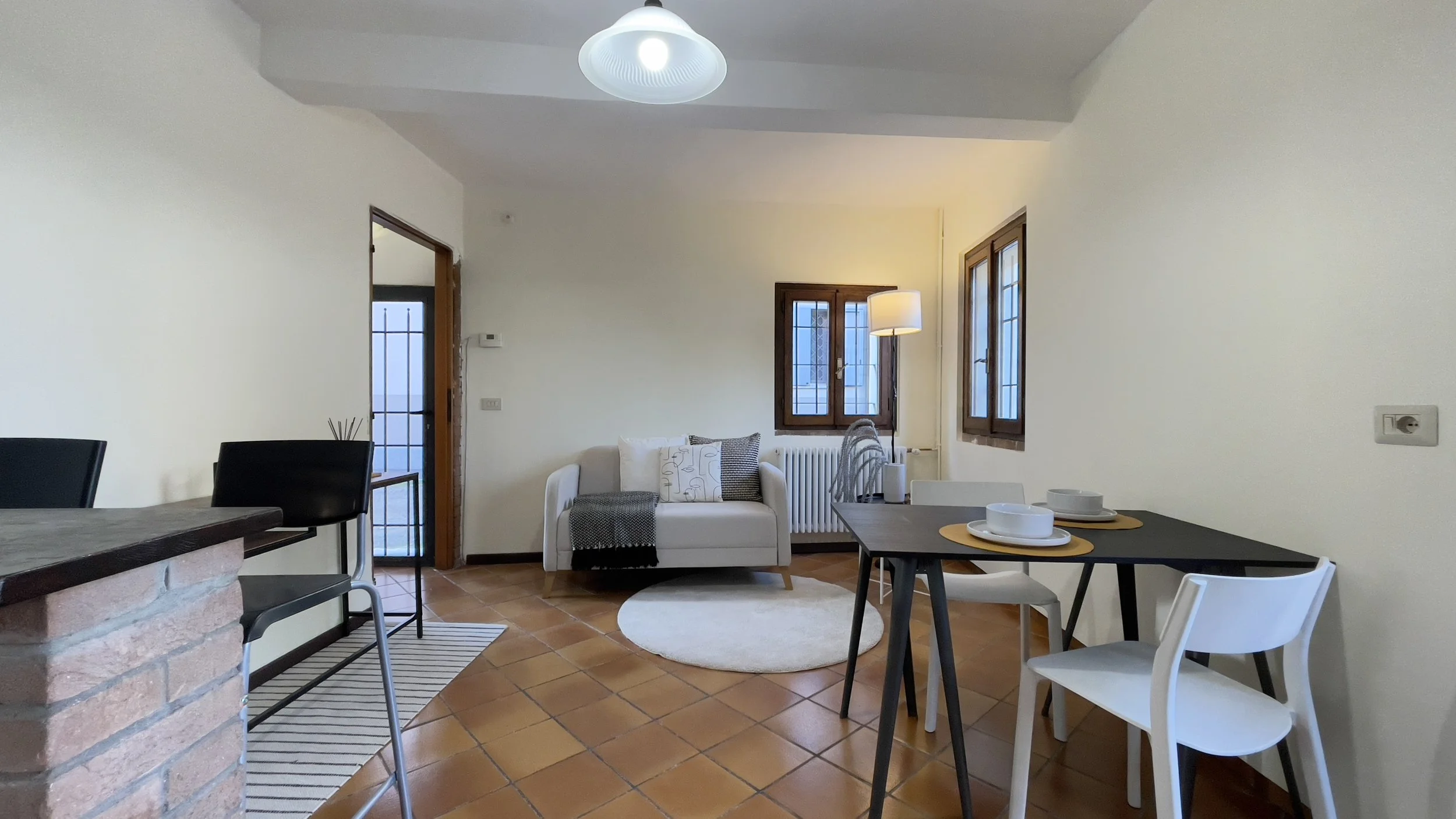 Soggiorno moderno con divano bianco, tavolo da pranzo nero, sedie bianche, lampada da terra, e finestre con grate di sicurezza.