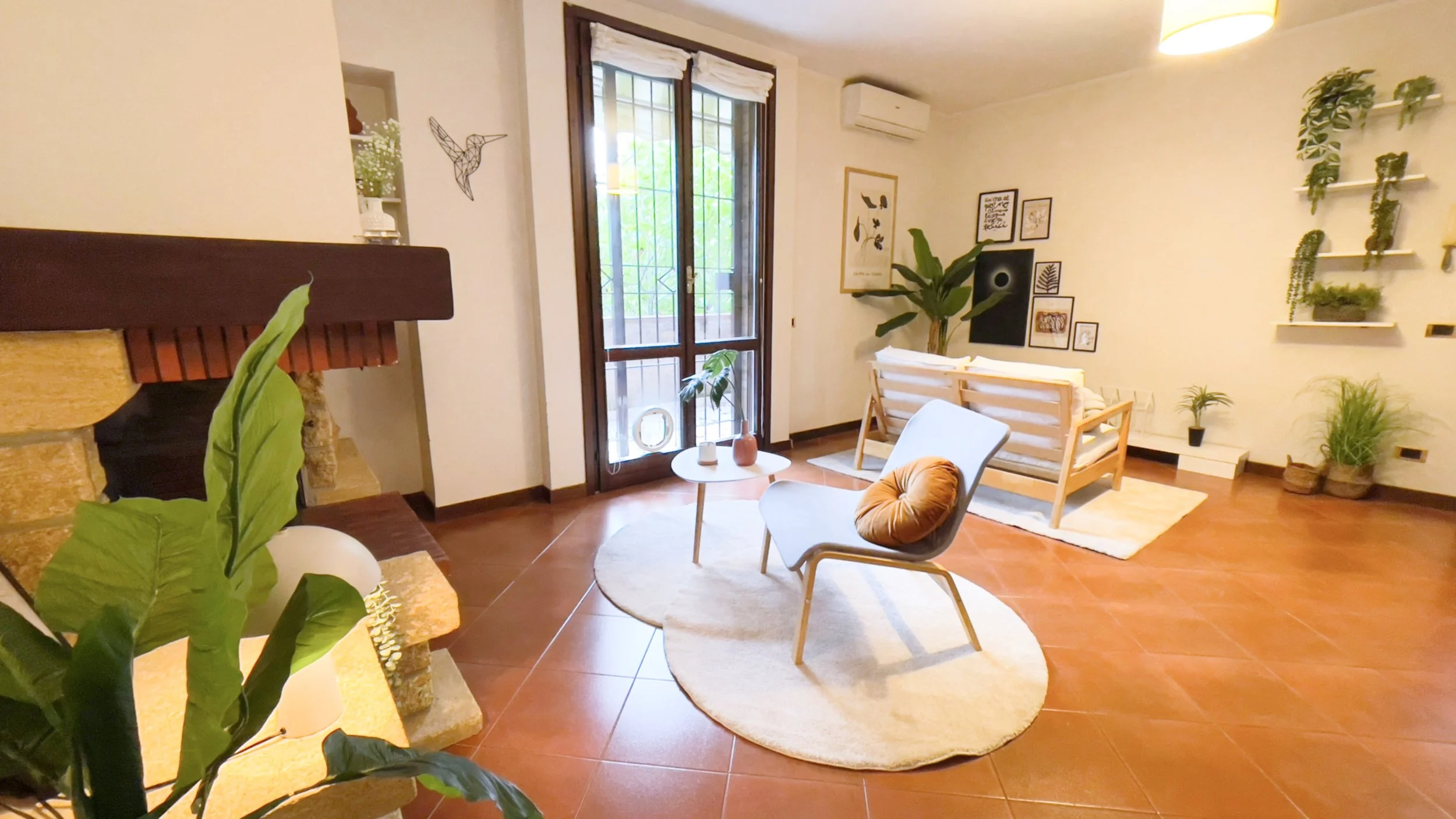 Soggiorno moderno con camino, divano bianco, poltrona grigia con cuscino arancione, tavolino rotondo bianco, piante decorative e parete con quadri e mensole. Pavimento in terracotta e ampia finestra con tende.