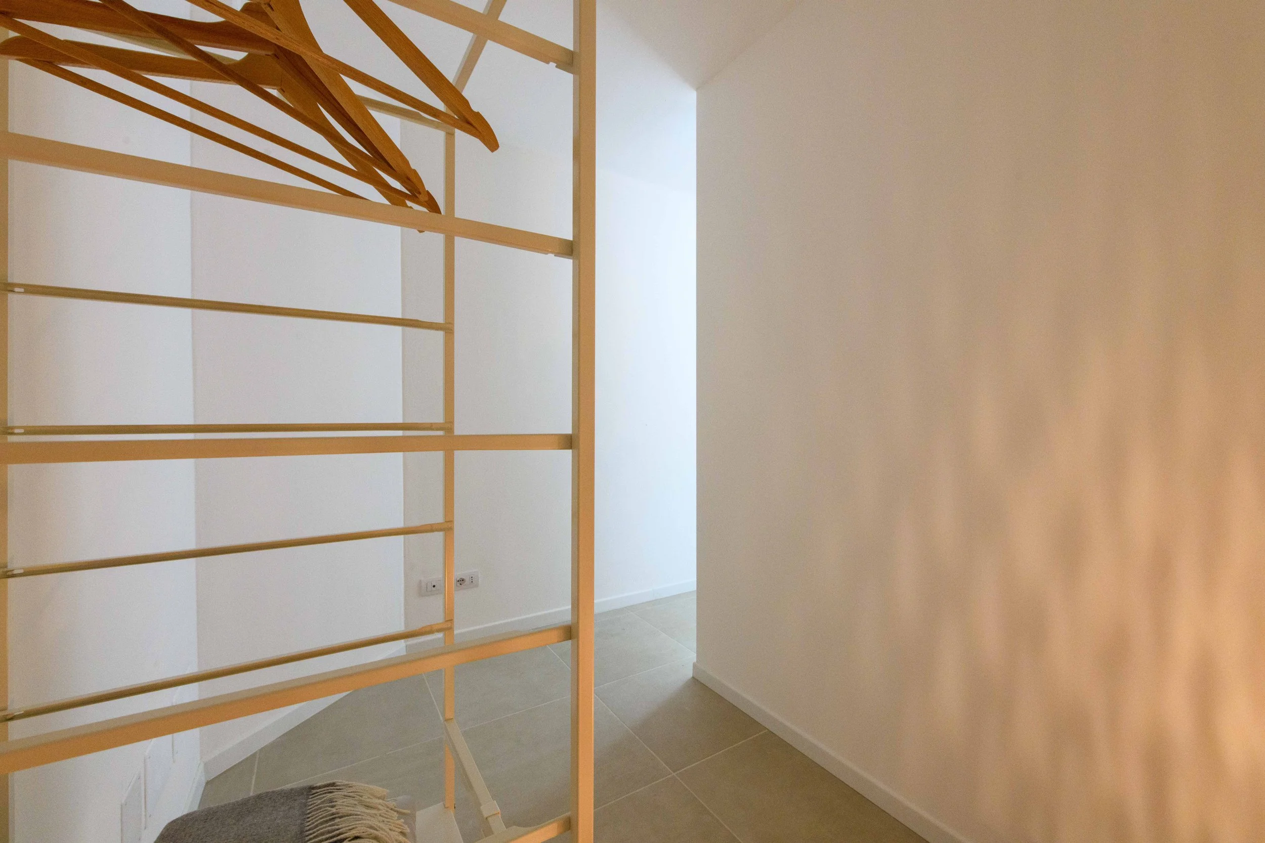 Interno di una stanza minimalista con pareti bianche, un appendiabiti di legno chiaro e un pavimento in piastrelle color beige chiaro. Ci sono delle grucce appese al supporto.