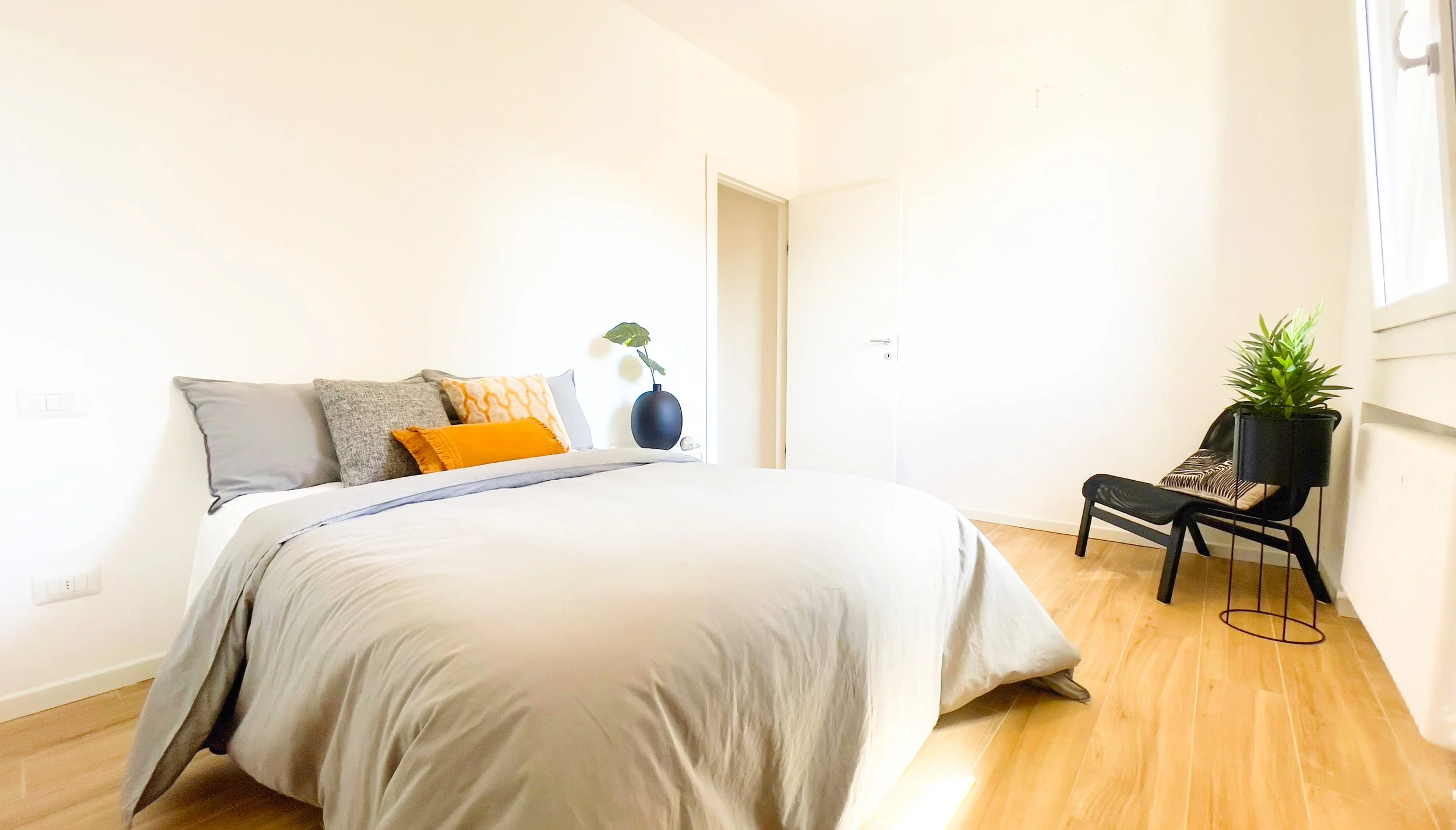 Camera da letto moderna con letto matrimoniale, cuscini colorati, pianta su vaso nero, sedia con pianta, pavimento in legno.