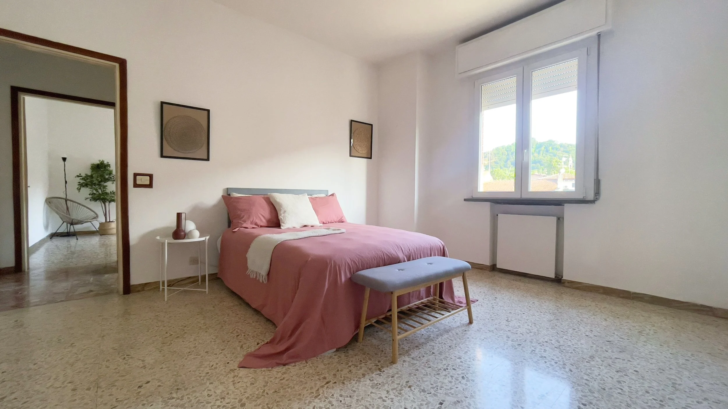 Camera da letto con letto matrimoniale con copriletto rosa, cuscini e coperta bianca. Finestra con vista esterna, pavimento in marmo chiaro e panca ai piedi del letto. Tavolino con decorazioni e cornici alle pareti. Vista su altra stanza con sedia e 