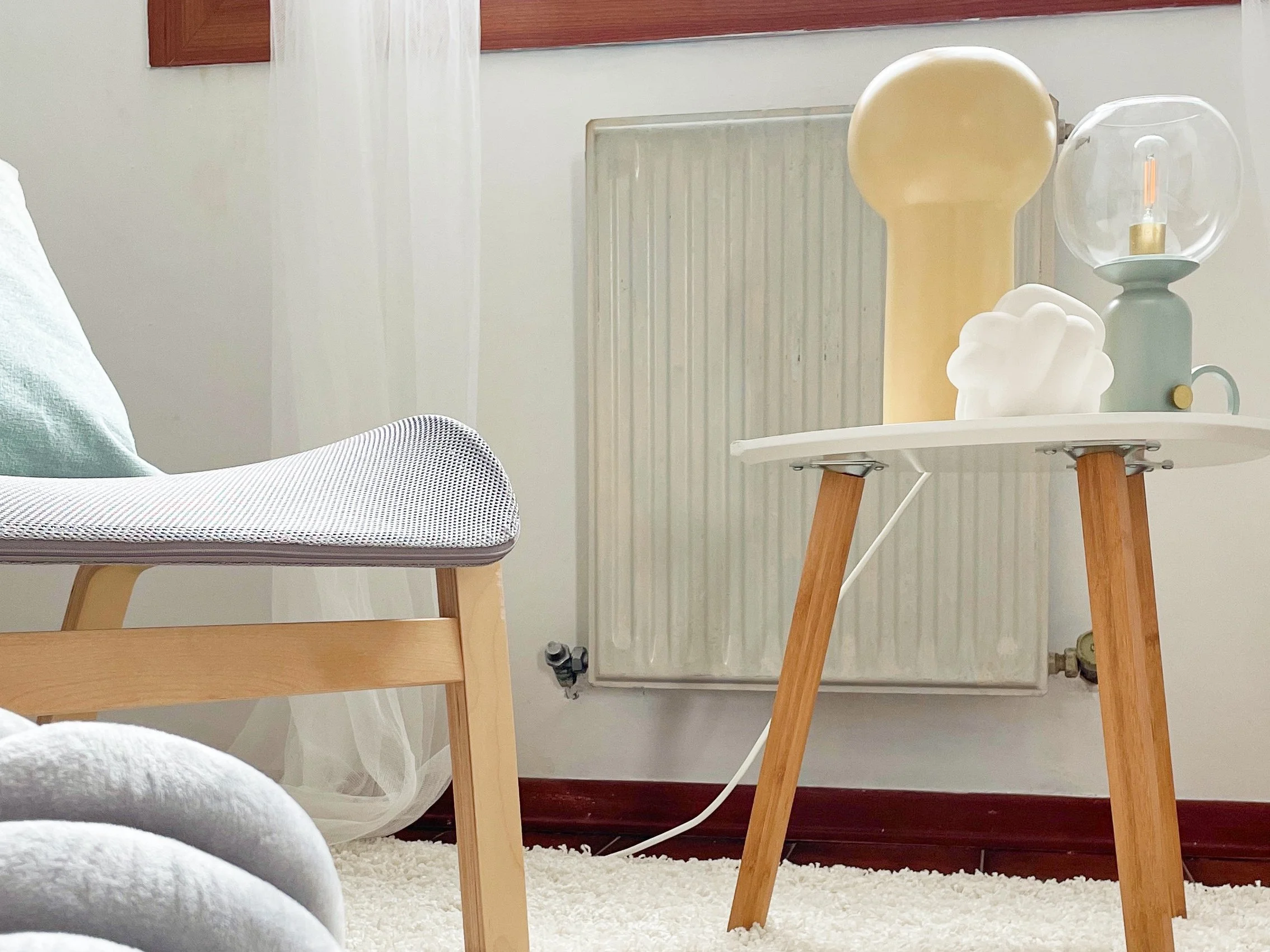 Interno di una stanza con sedia moderna e tavolino con lampada e decorazione, pavimento con tappeto bianco, sfondo con termosifone e tenda.