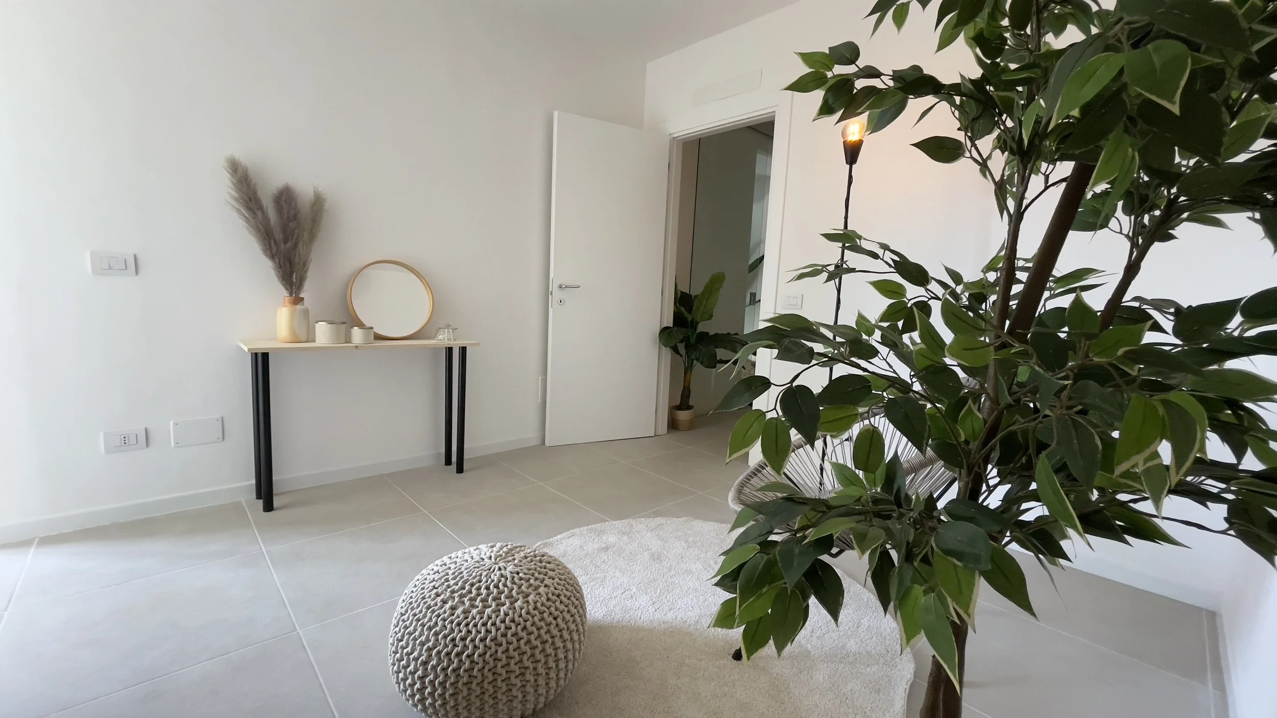 Stanza minimalista con pianta, tavolino con specchio e decorazioni, pouf intrecciato, tappeto chiaro e porta aperta.