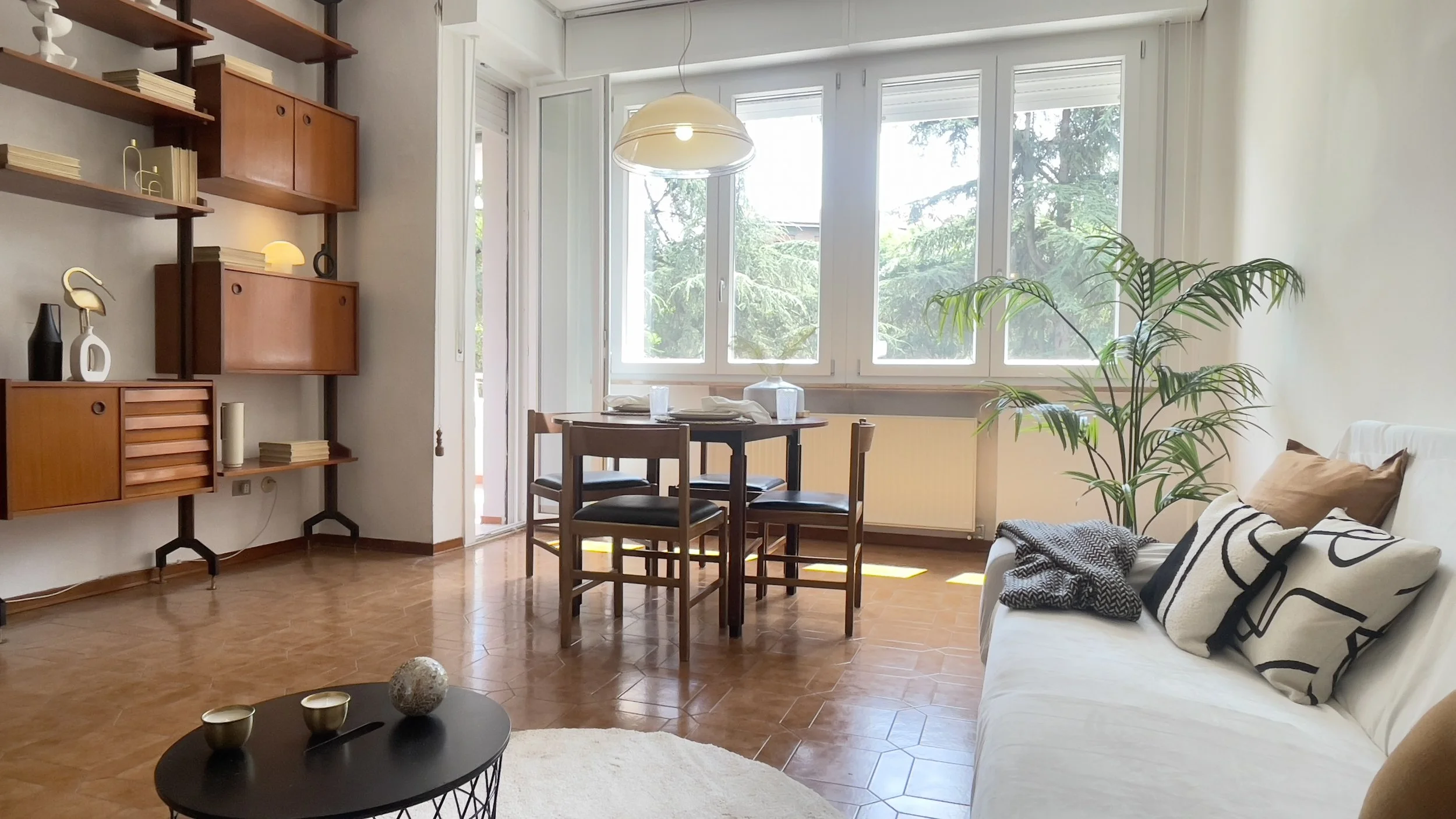 Soggiorno luminoso con tavolo da pranzo e sedie, libreria a muro, divano con cuscini decorativi, pianta verde e grandi finestre che lasciano entrare luce naturale.