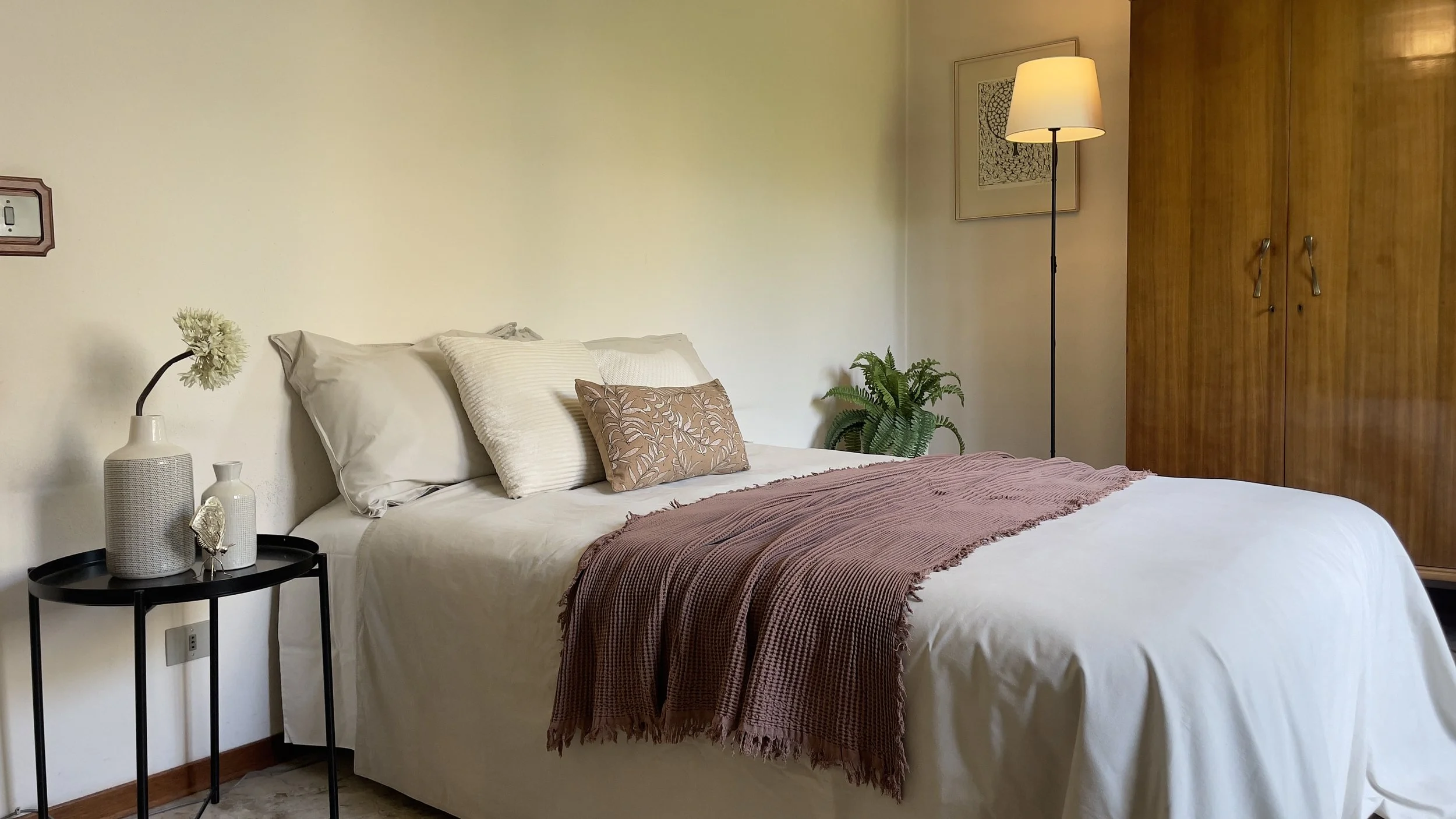 Camera da letto moderna con letto matrimoniale, coperte bianche, cuscini decorativi, lampada da terra, armadio di legno e piante decorative.