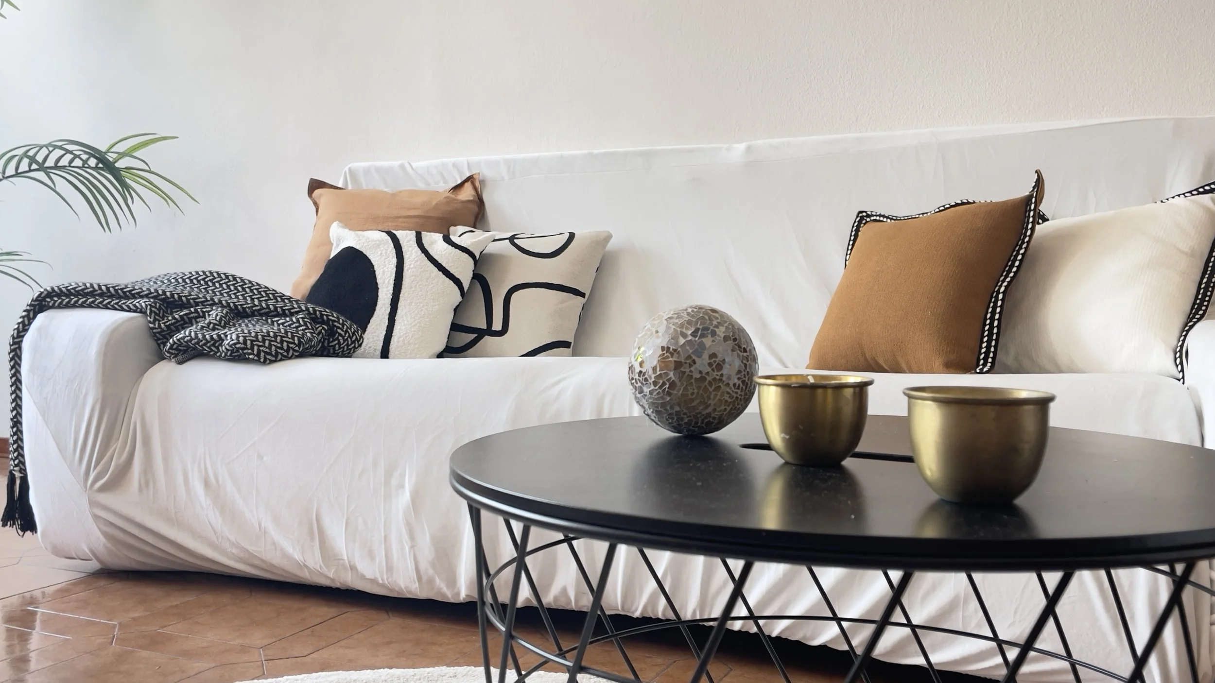 Divano bianco con cuscini colorati, coperta a motivi geometrici, tavolino nero con decorazione sferica e due tazze dorate, pianta sulla sinistra.