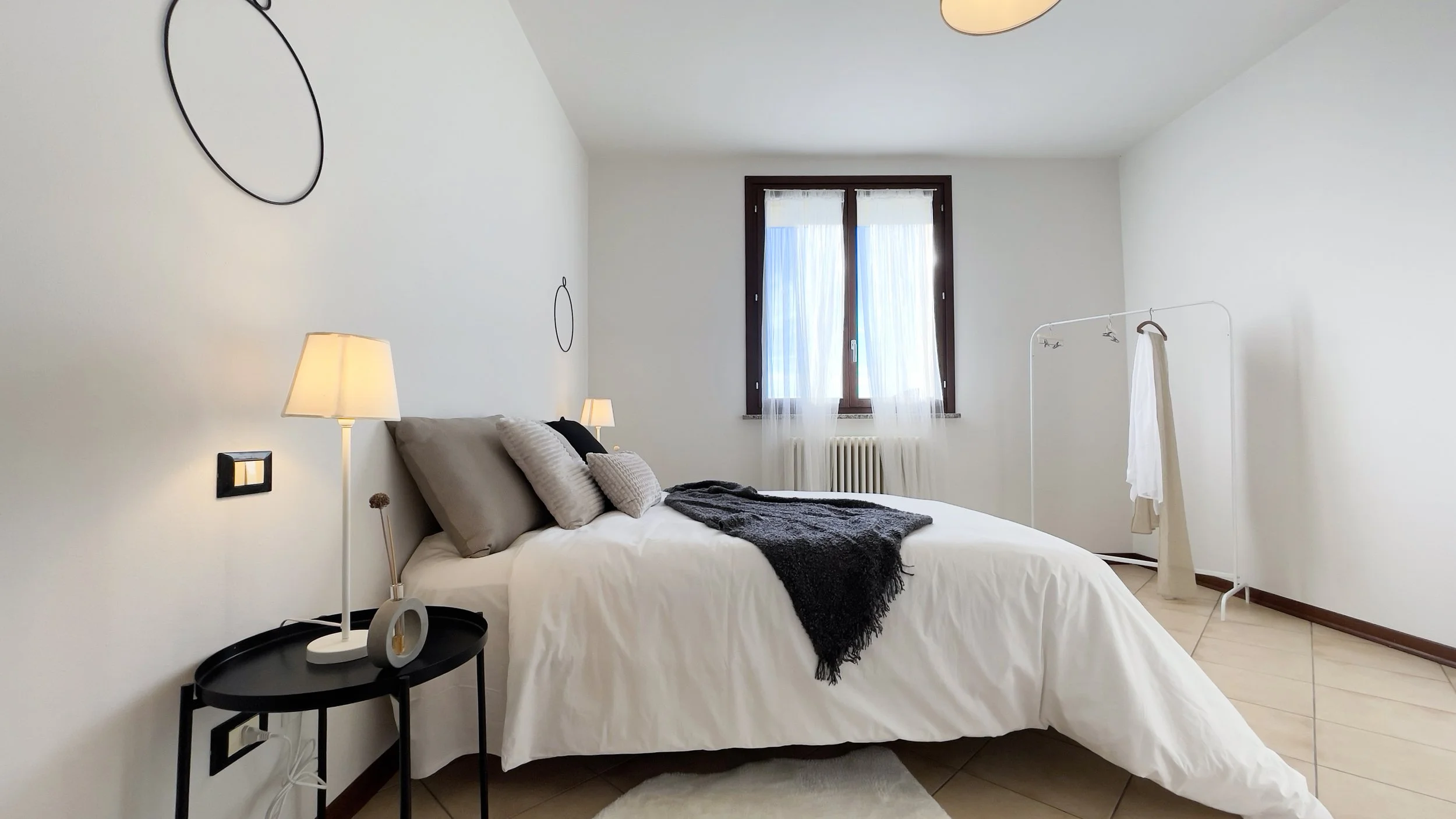 Camera da letto moderna con letto matrimoniale, cuscini grigi e bianchi, coperta scura, lampada da tavolo su comodino nero, specchio rotondo sul muro, finestra con tende bianche, appendiabiti bianco e pavimento piastrellato beige.