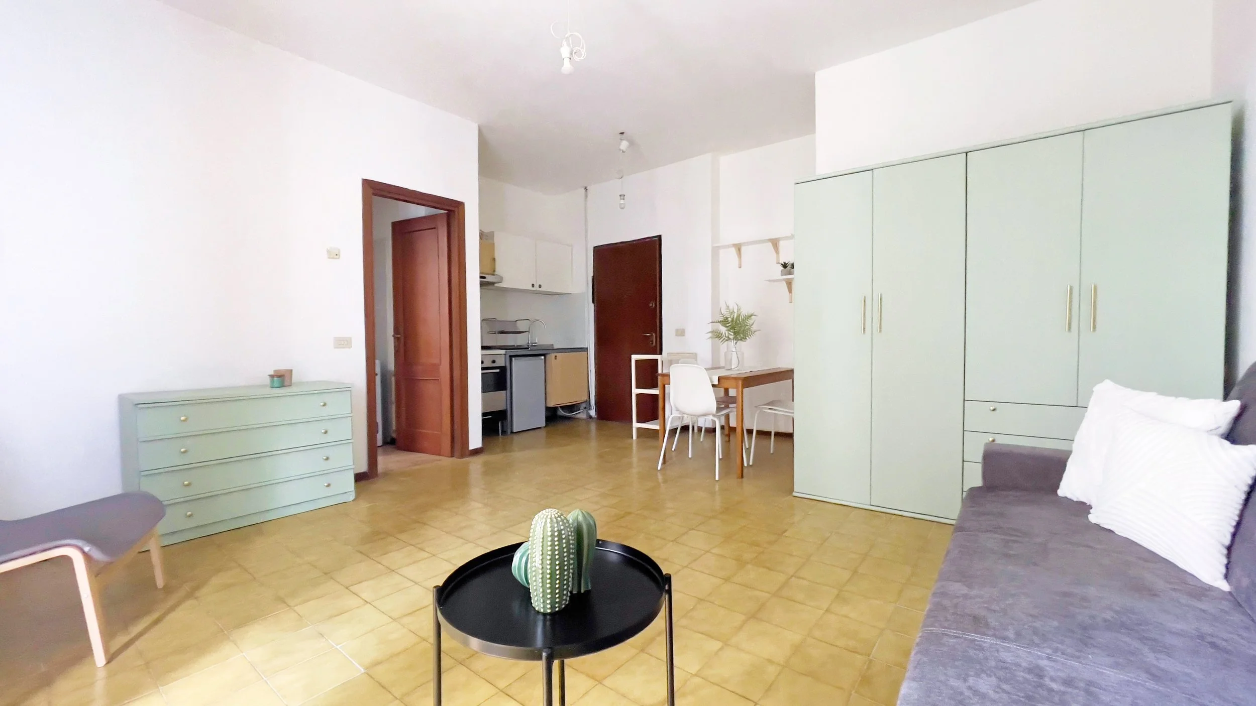 Interno di un appartamento con mobili moderni. A sinistra un comò verde chiaro con candele sopra. Sul pavimento c'è un tavolino nero con un cactus decorativo. Sul lato destro c'è un armadio verde chiaro e un divano grigio con cuscini bianchi. In fond