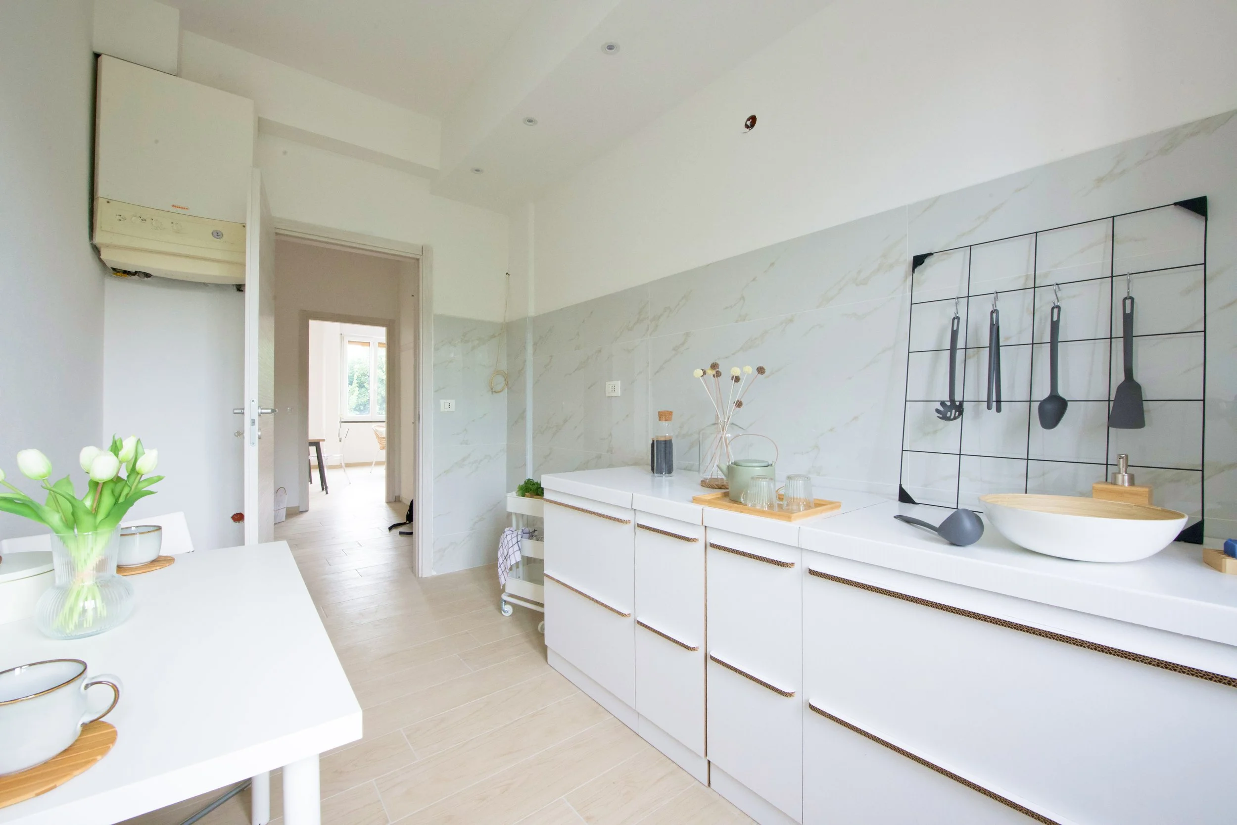 Cucina moderna luminosa con mobili bianchi, decorazioni minimaliste, piani d'appoggio chiari e utensili appesi alla parete. Tavolo con fiori bianchi e tazze sulla sinistra.
