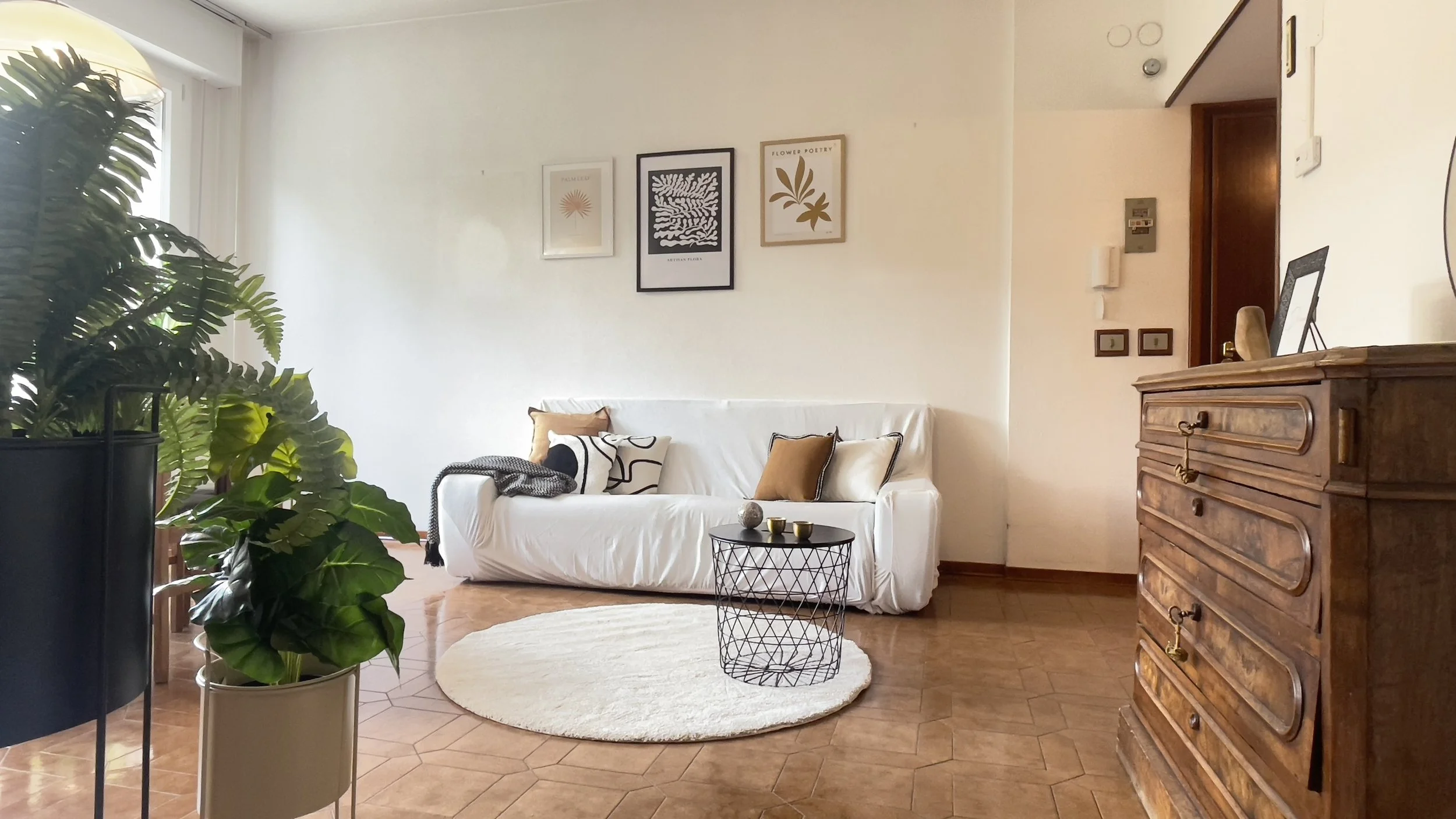 Salotto moderno con divano bianco, piante in vasi, tavolo rotondo nero su tappeto chiaro, e un mobile in legno.