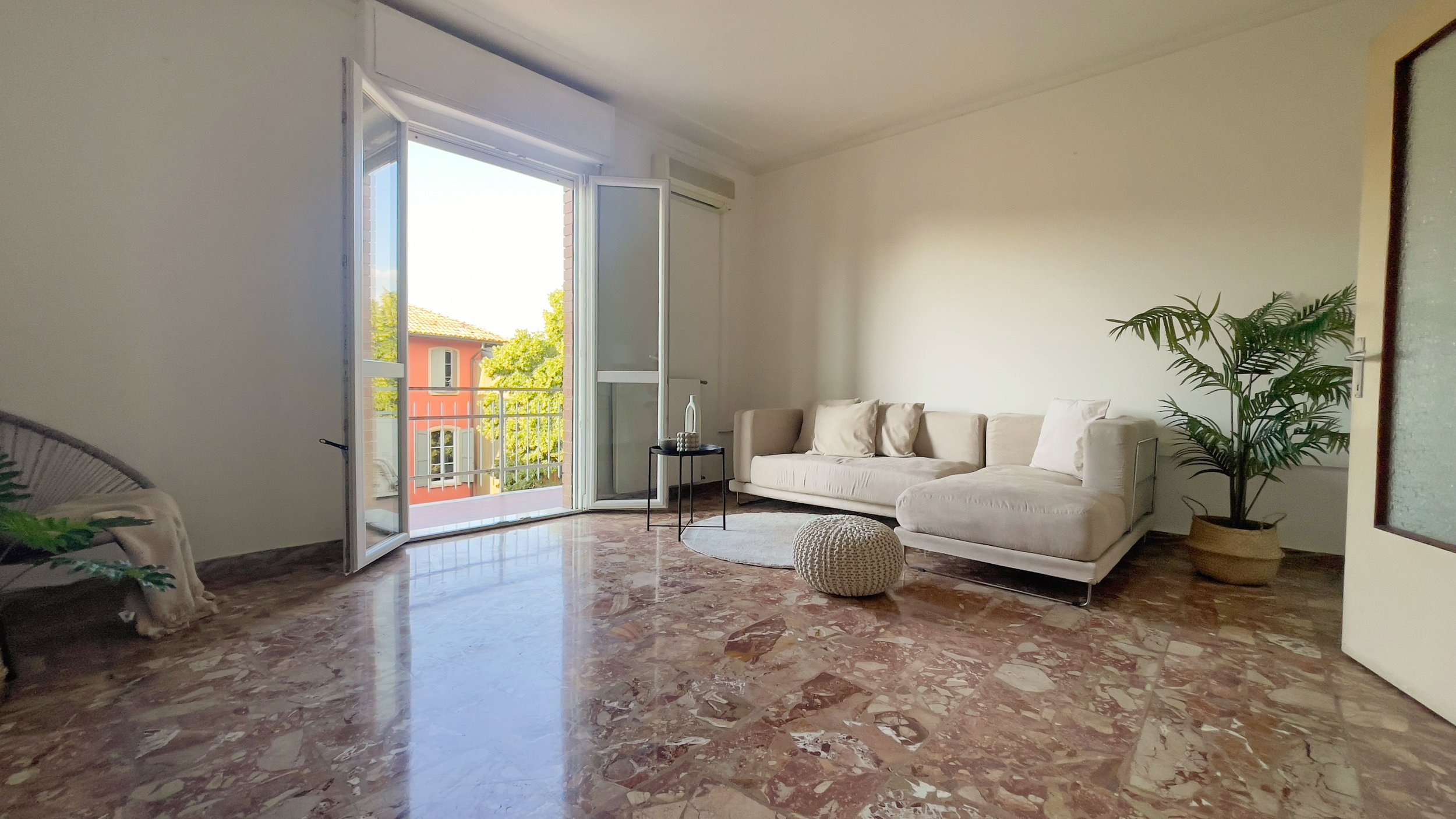 Salotto moderno con divano beige, tavolino rotondo nero, cuscino a maglia e pianta decorativa. Pavimento in marmo lucido. Porta-finestra aperta su balcone con vista su edificio arancione.