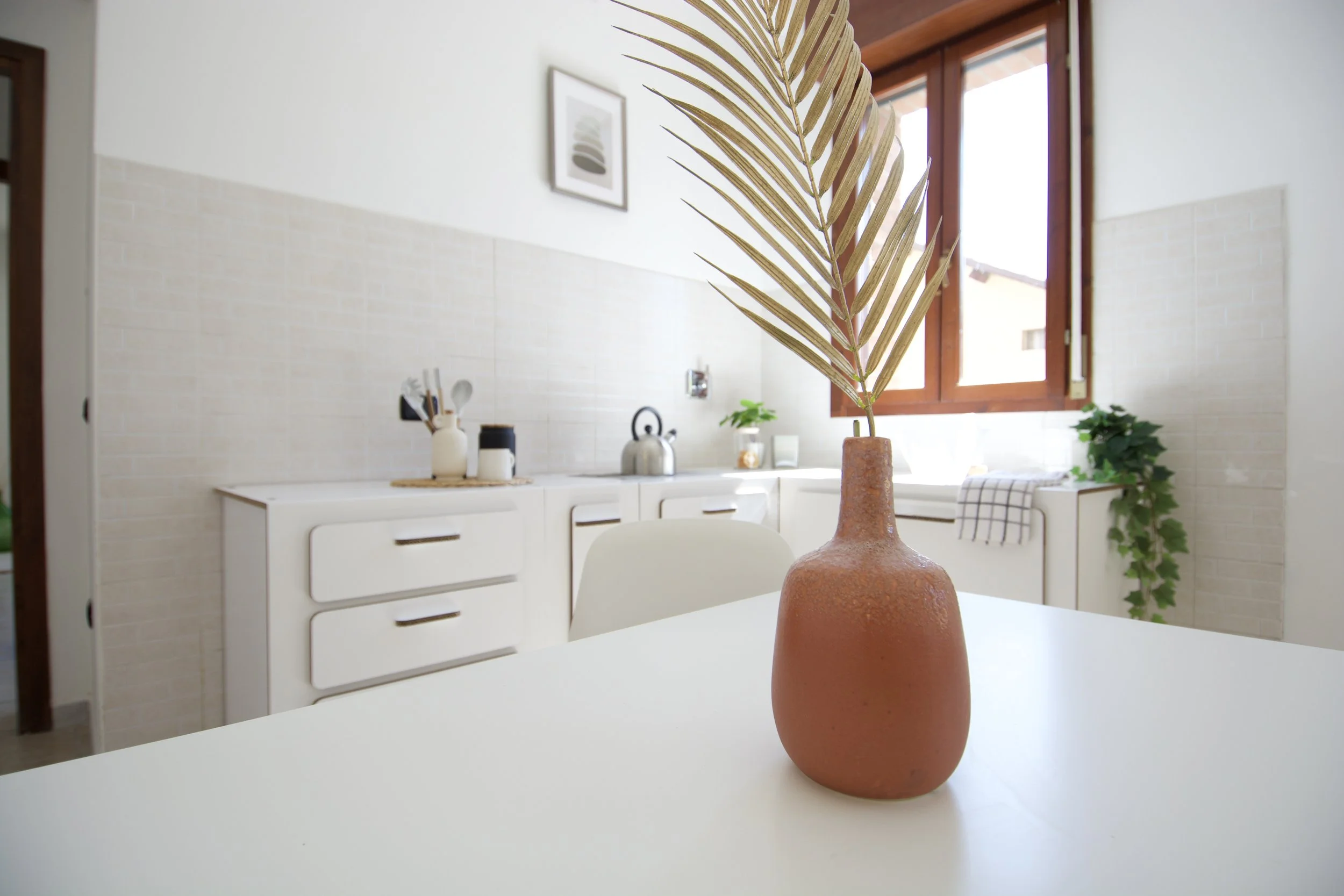 Cucina moderna con vaso di ceramica e foglia secca su tavolo bianco, finestre e mobili bianchi sullo sfondo.