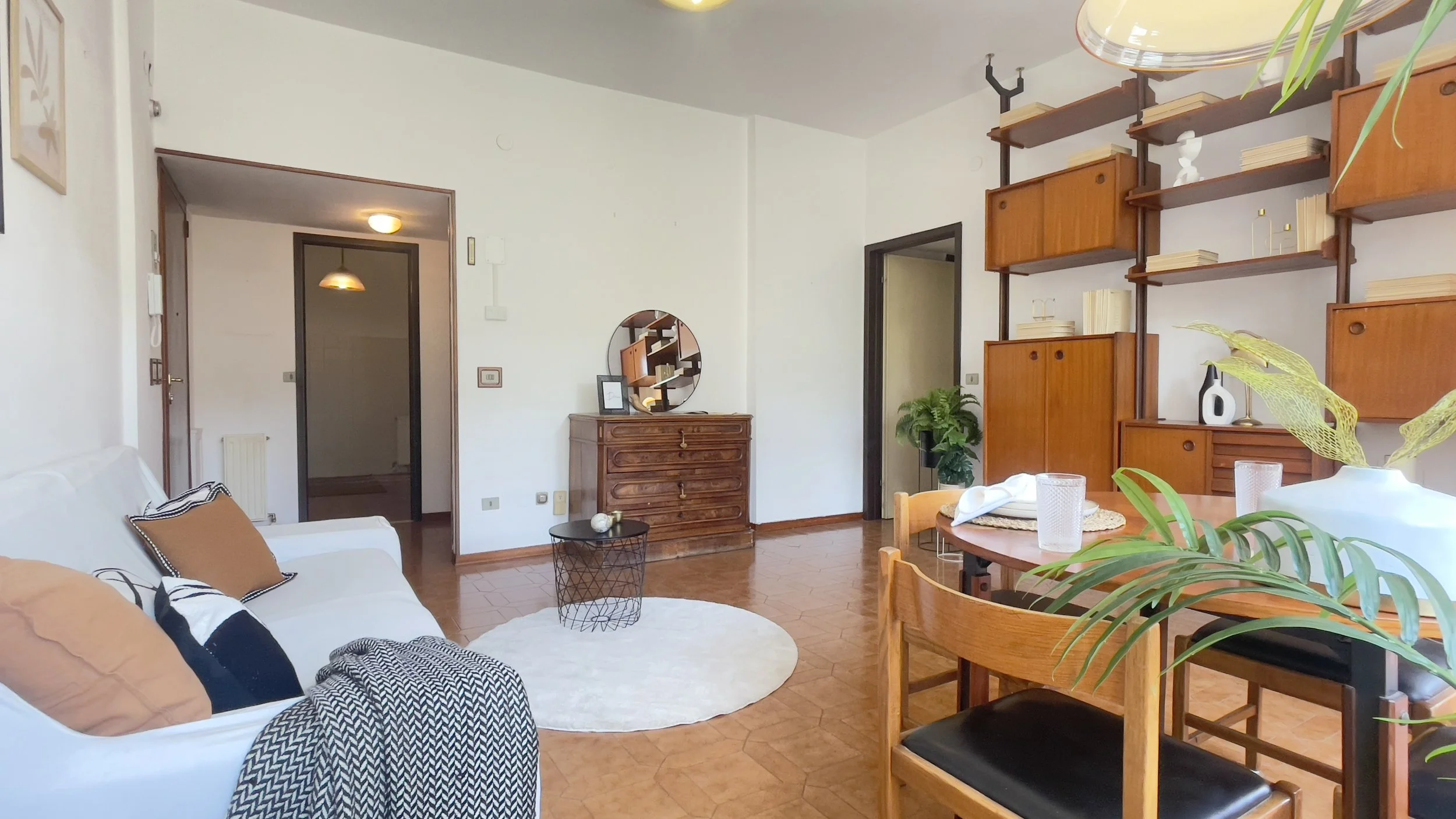 Soggiorno moderno con divano bianco, cuscini colorati e plaid. Mobile con specchio rotondo, tavolino nero e tappeto rotondo bianco. Scaffalatura in legno con libri e decorazioni, pianta verde e tavolo da pranzo con sedie in legno.