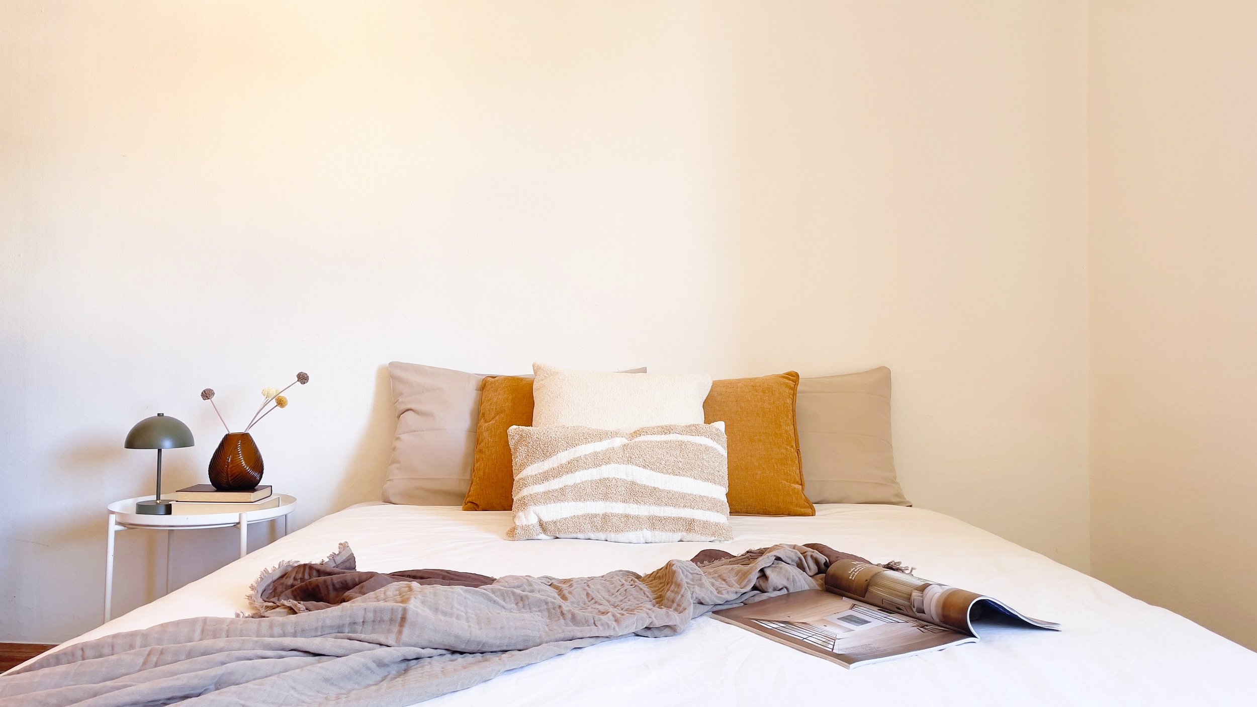 Camera da letto minimalista con letto matrimoniale, cuscini decorativi, coperta e tavolino con lampada e vaso.