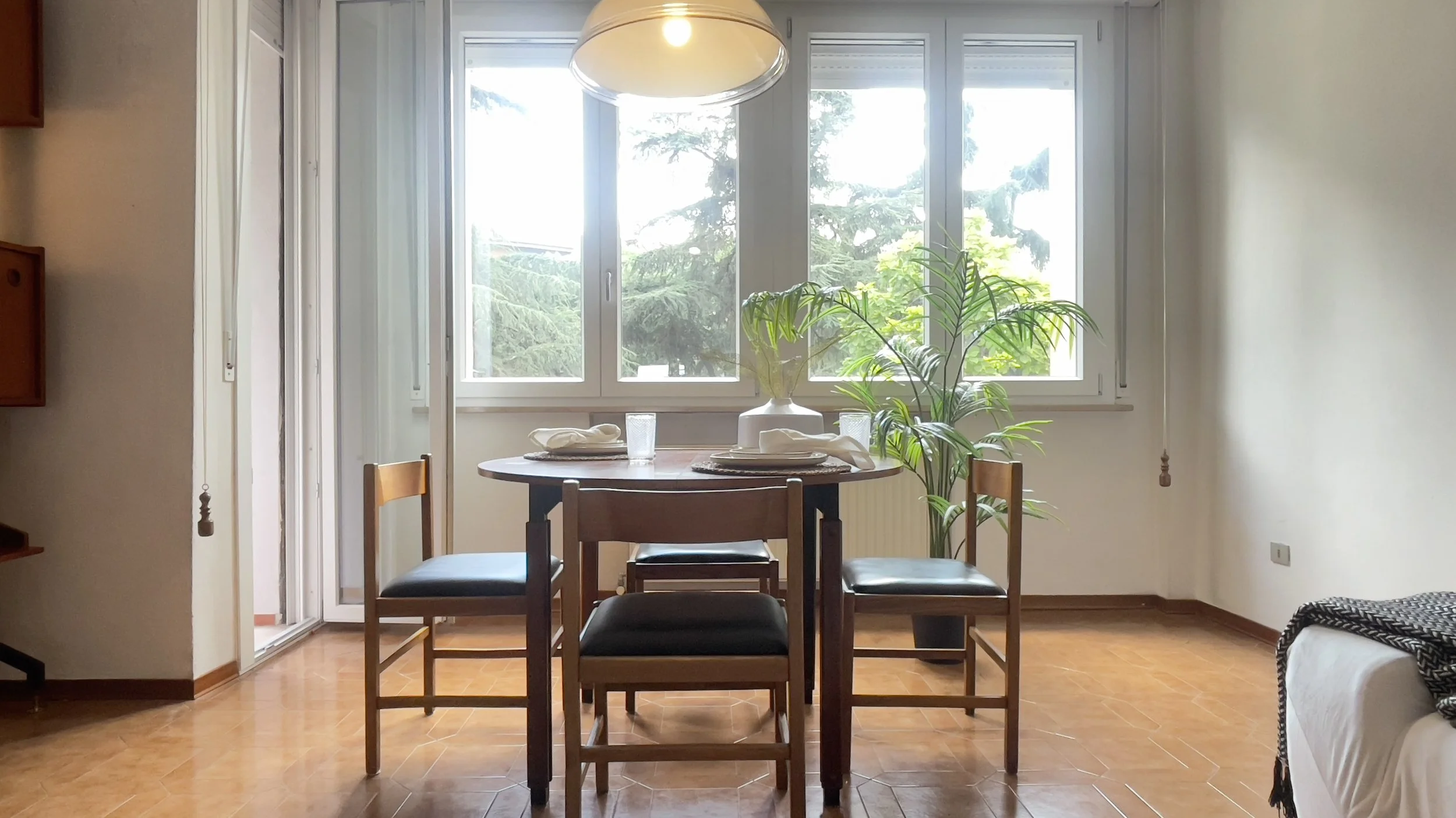 Sala da pranzo con tavolo rotondo e quattro sedie, pianta interna vicino alla finestra, luce pendente sopra il tavolo, pavimento in legno.