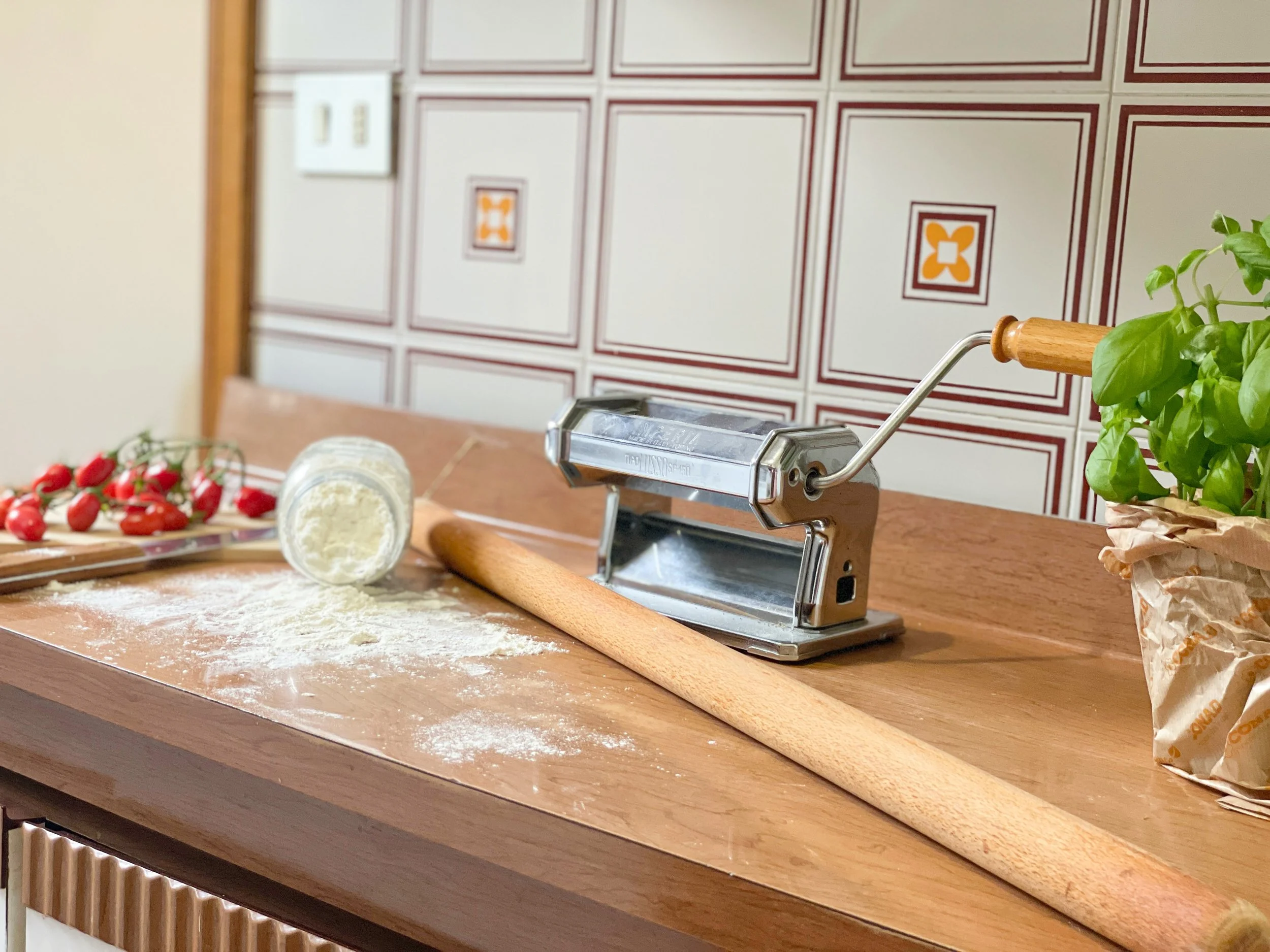 Macchina per pasta su un tavolo in cucina con farina, mattarello, pomodori e vaso di basilico.