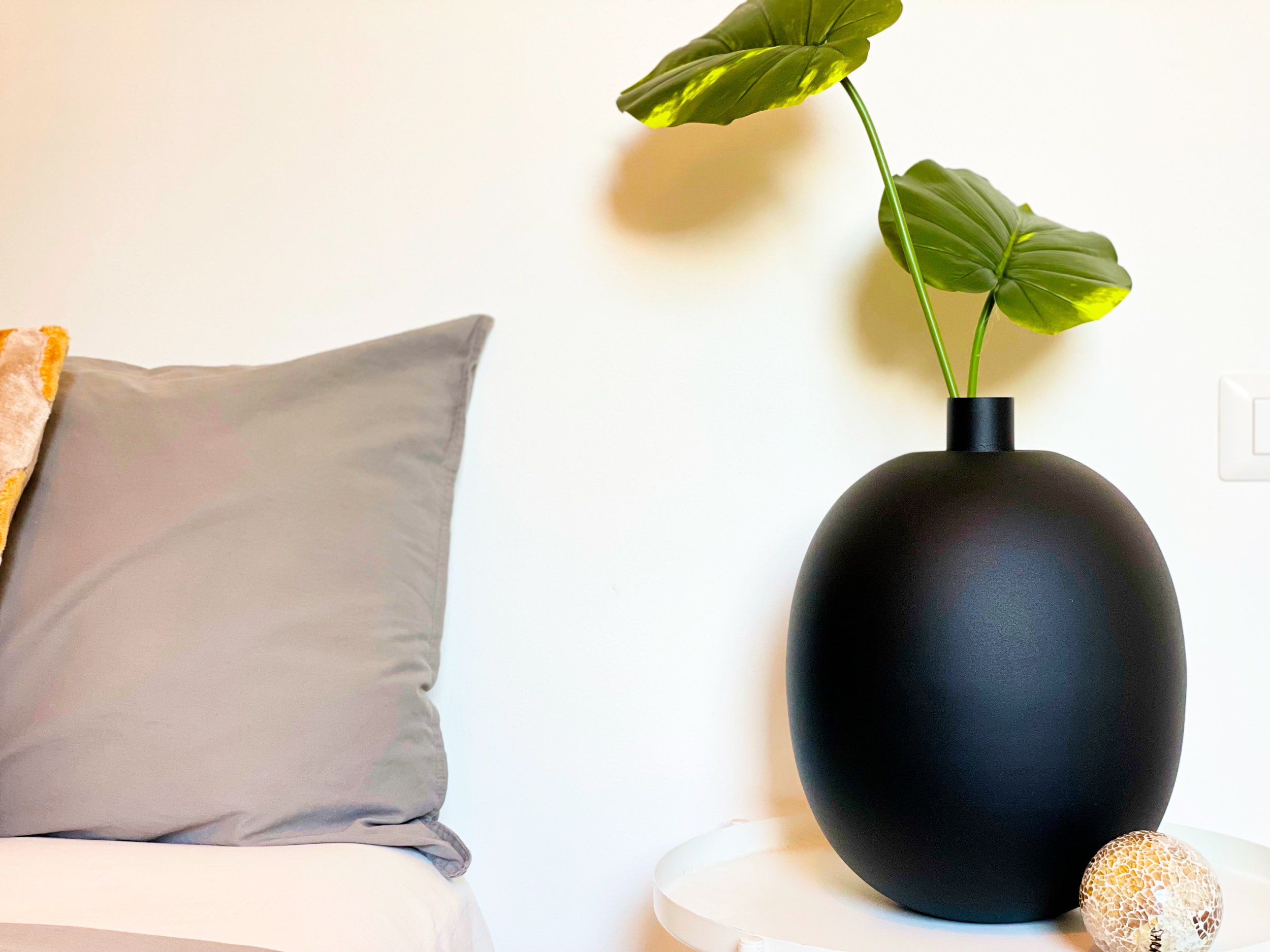 Pianta in vaso nero su tavolino accanto a letto con cuscino grigio.