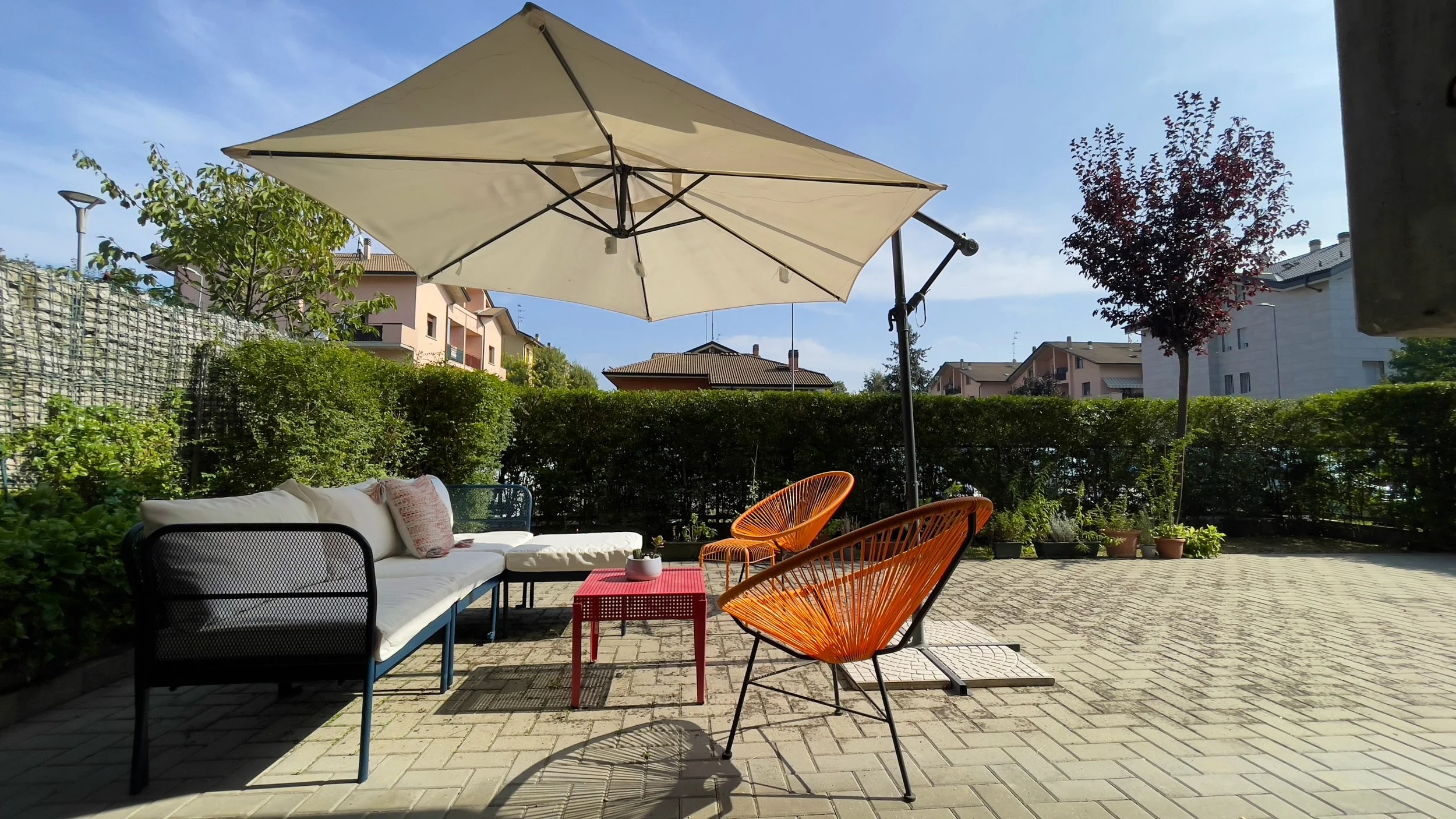 Area esterna con poltrone colorate, tavolino rosso e ombrellone, circondata da siepi e alberi, cielo sereno.