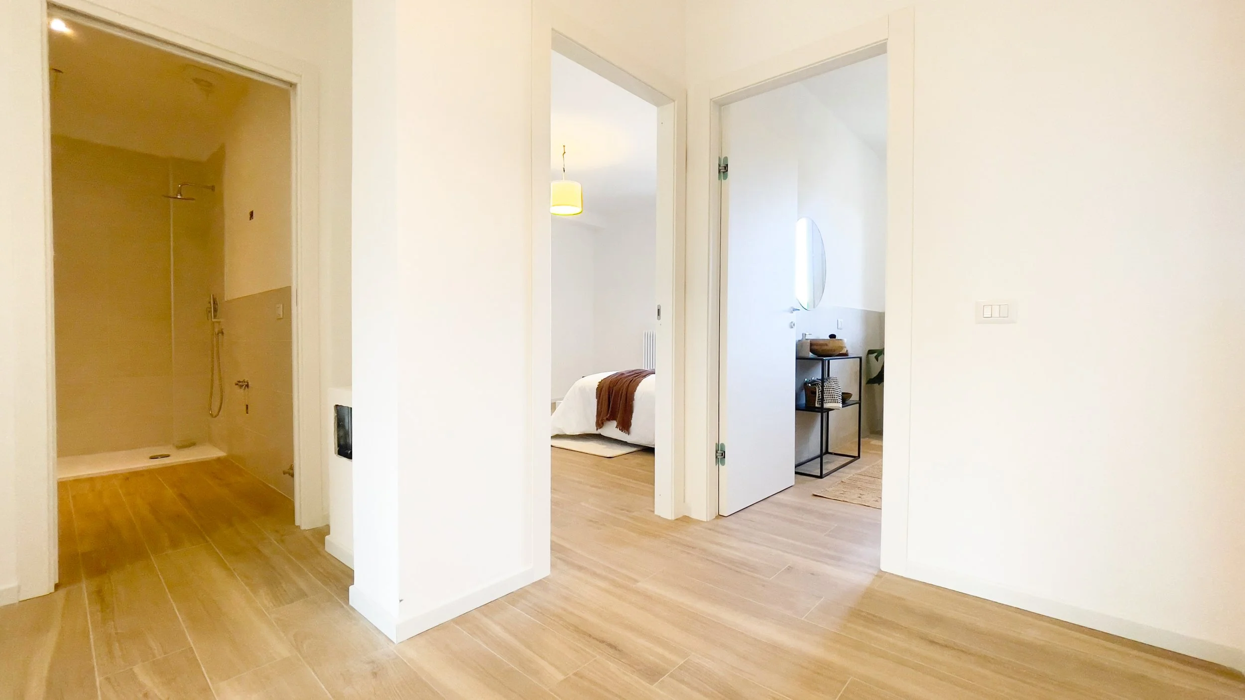 Interno di una casa con pavimento in legno chiaro, un bagno visibile a sinistra e una camera da letto al centro. Il bagno ha una doccia con piastrelle beige. La camera da letto ha un letto con coperta marrone. A destra c'è un angolo con uno specchio 