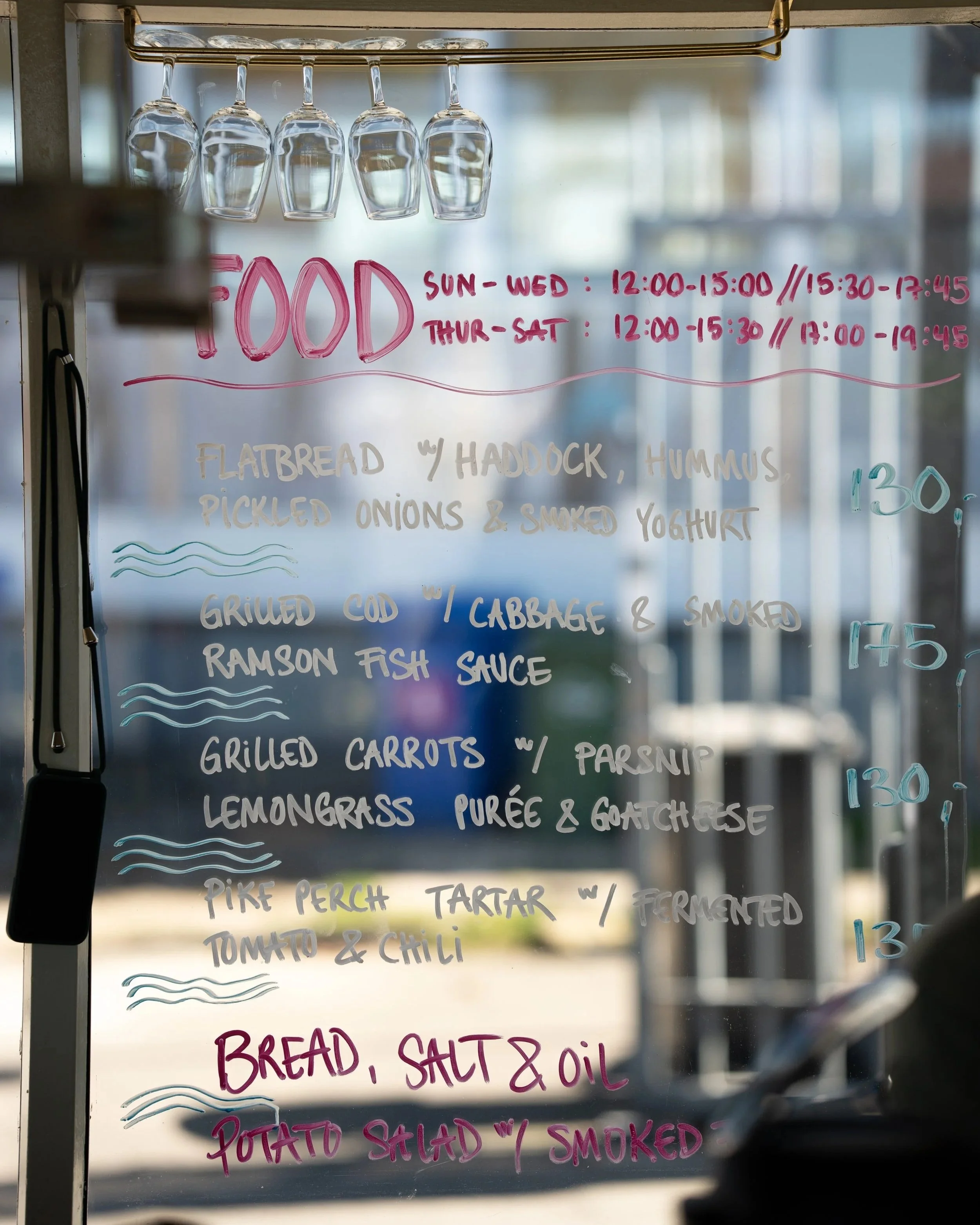 A handwritten menu on a glass window listing food items and their prices, with glassware hanging above. The menu includes hours of operation, and items such as flatbread with hummus and pickled onions, grilled cod with cabbage and smoked ramson fish 