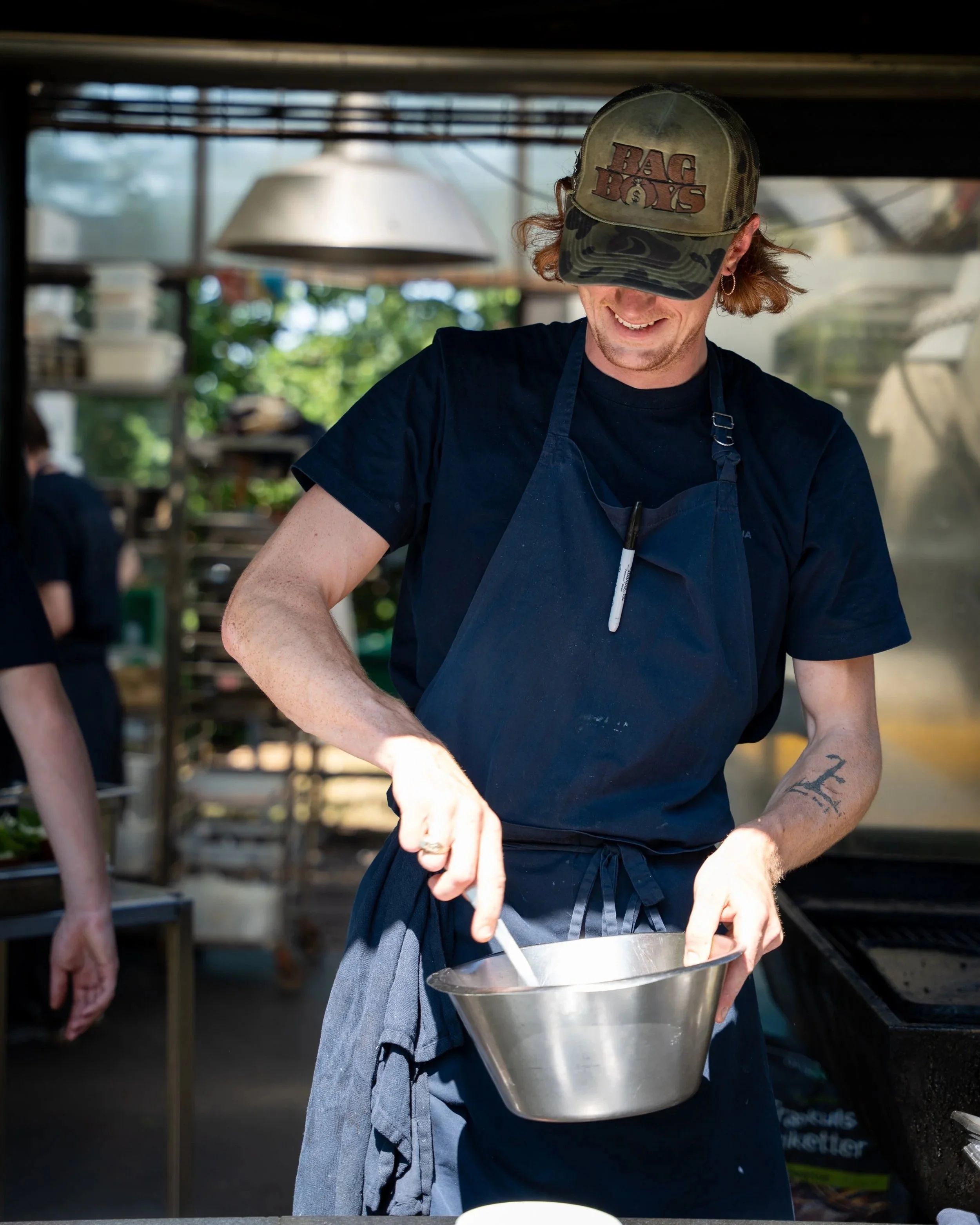 A person wearing a camouflage cap with the logo 'Bag & Bones' is smiling and holding a metal mixing bowl, with another person partially visible on the left. The background shows a blurred outdoor setting with trees and a metal structure overhead.