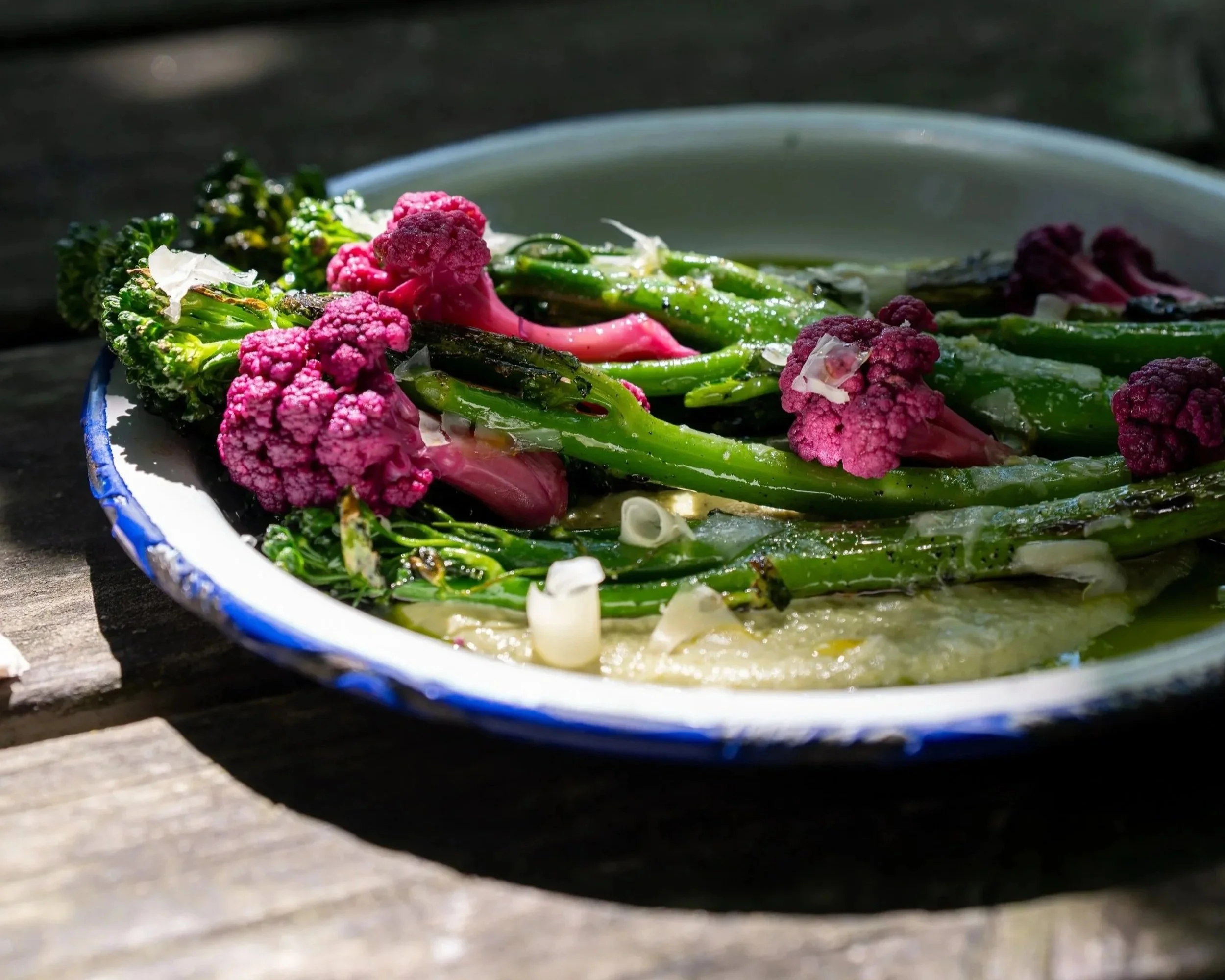 Close-up of colorful charred vegetables, including purple cauliflower and green leafy vegetables, on a white plate with a blue rim, placed on a wooden surface.