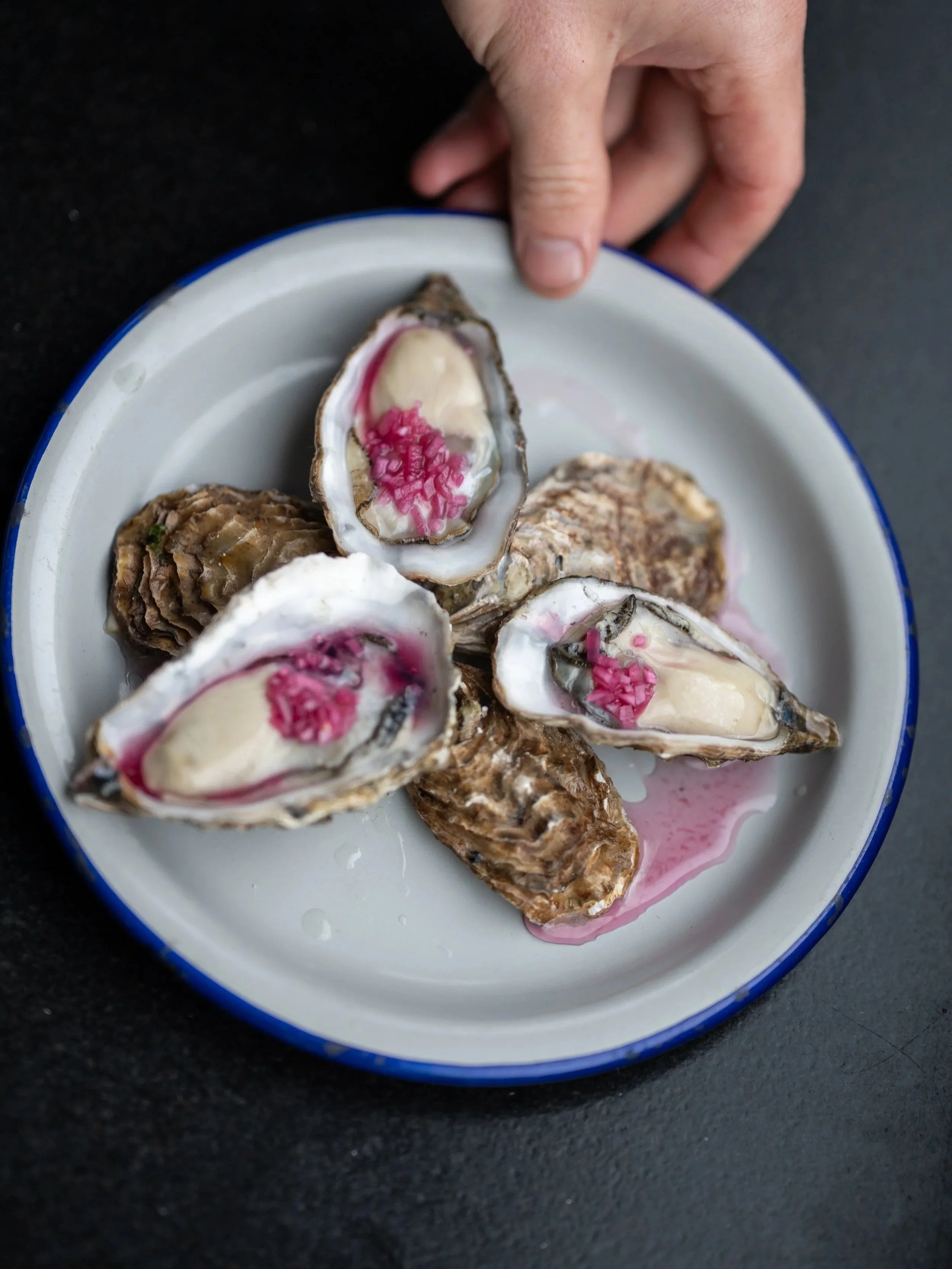 Close-up of an oyster with pink and white filling on a white plate with a blue rim, on a black surface.