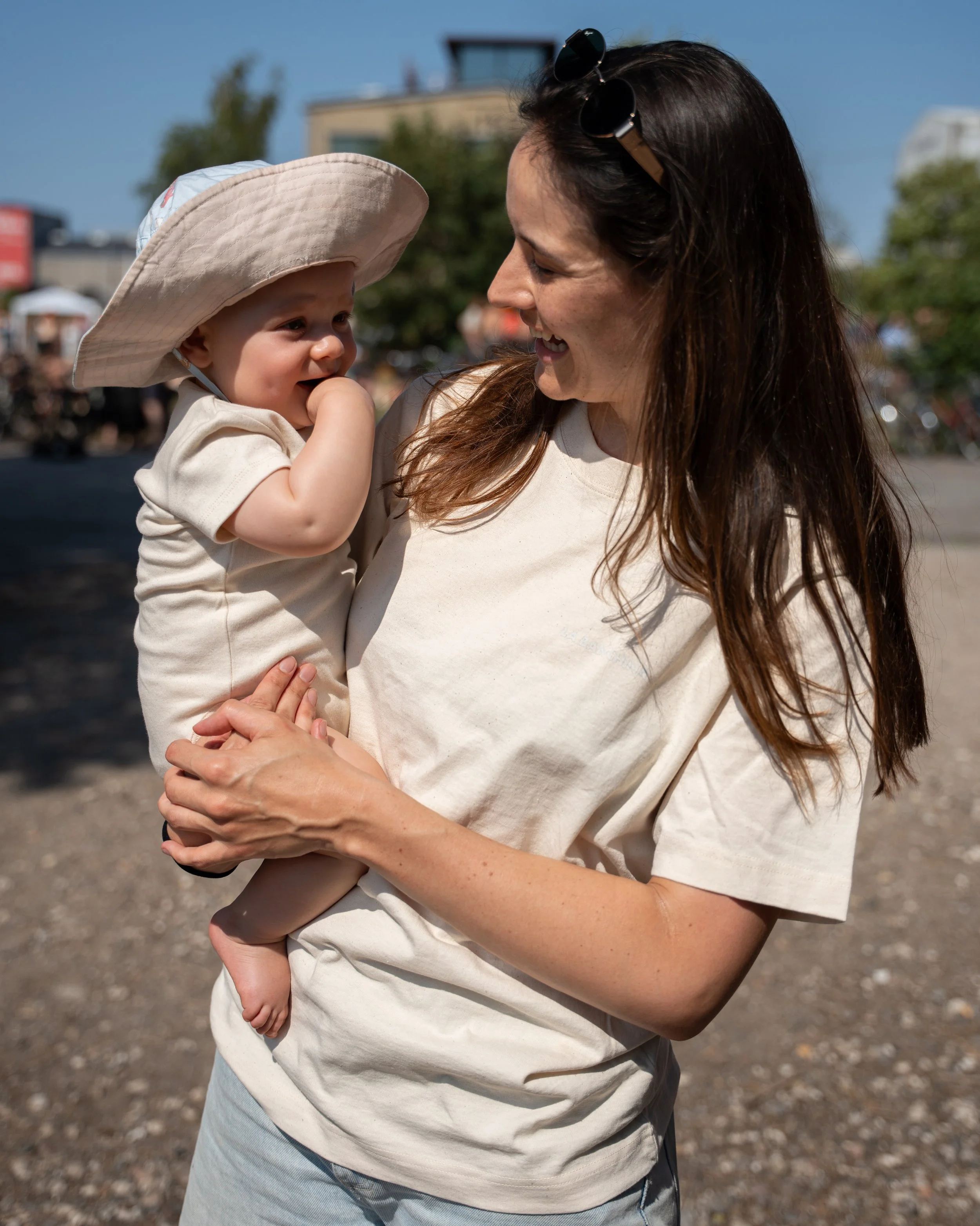A woman with long brown hair, wearing sunglasses on her head and a beige T-shirt, holding a smiling toddler wearing a large sun hat and a cream-colored T-shirt outdoors on a sunny day. The background shows a building and trees.